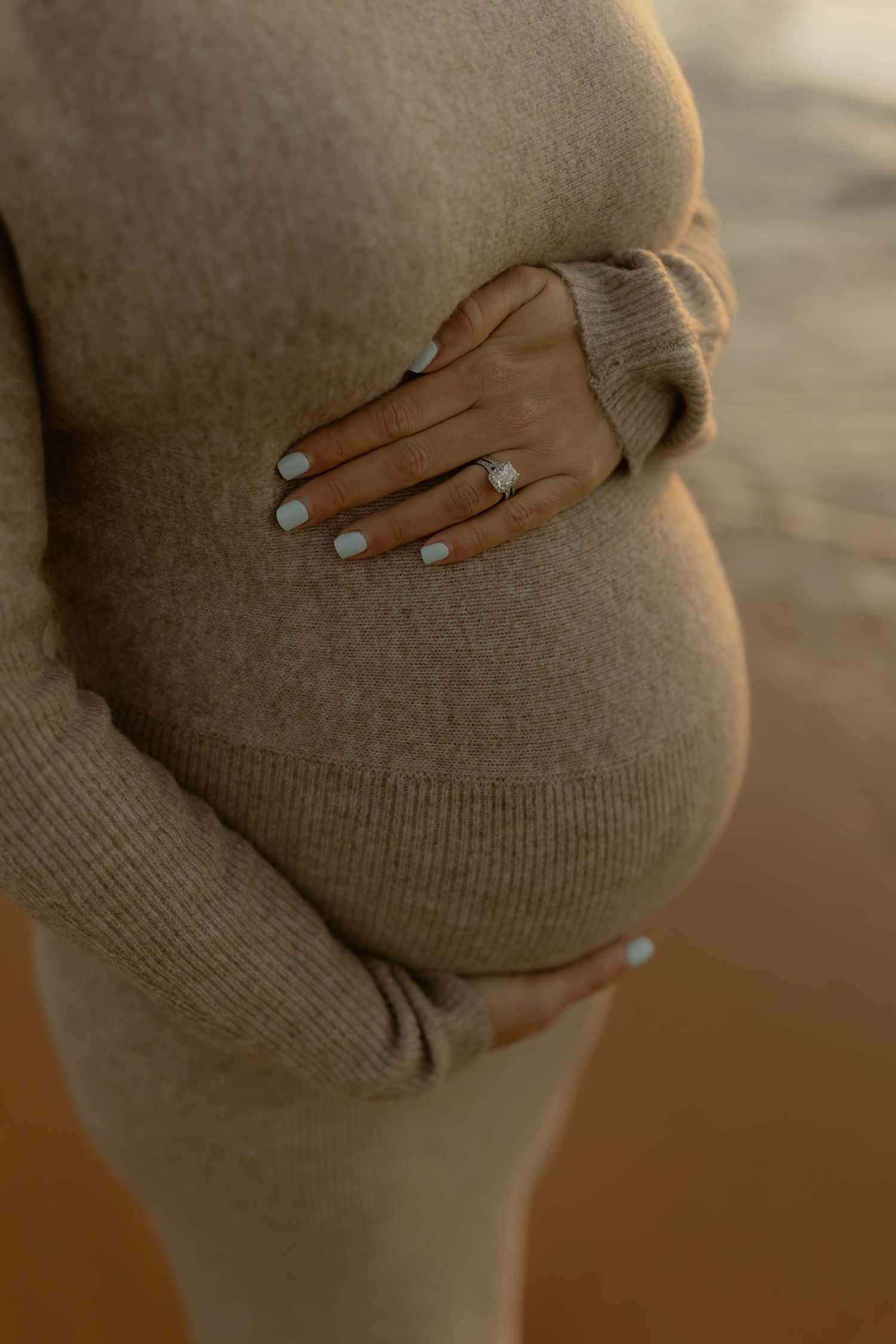 Close-up of pregnant woman in beige knit dress cradling baby bump with manicured hands and diamond ring.