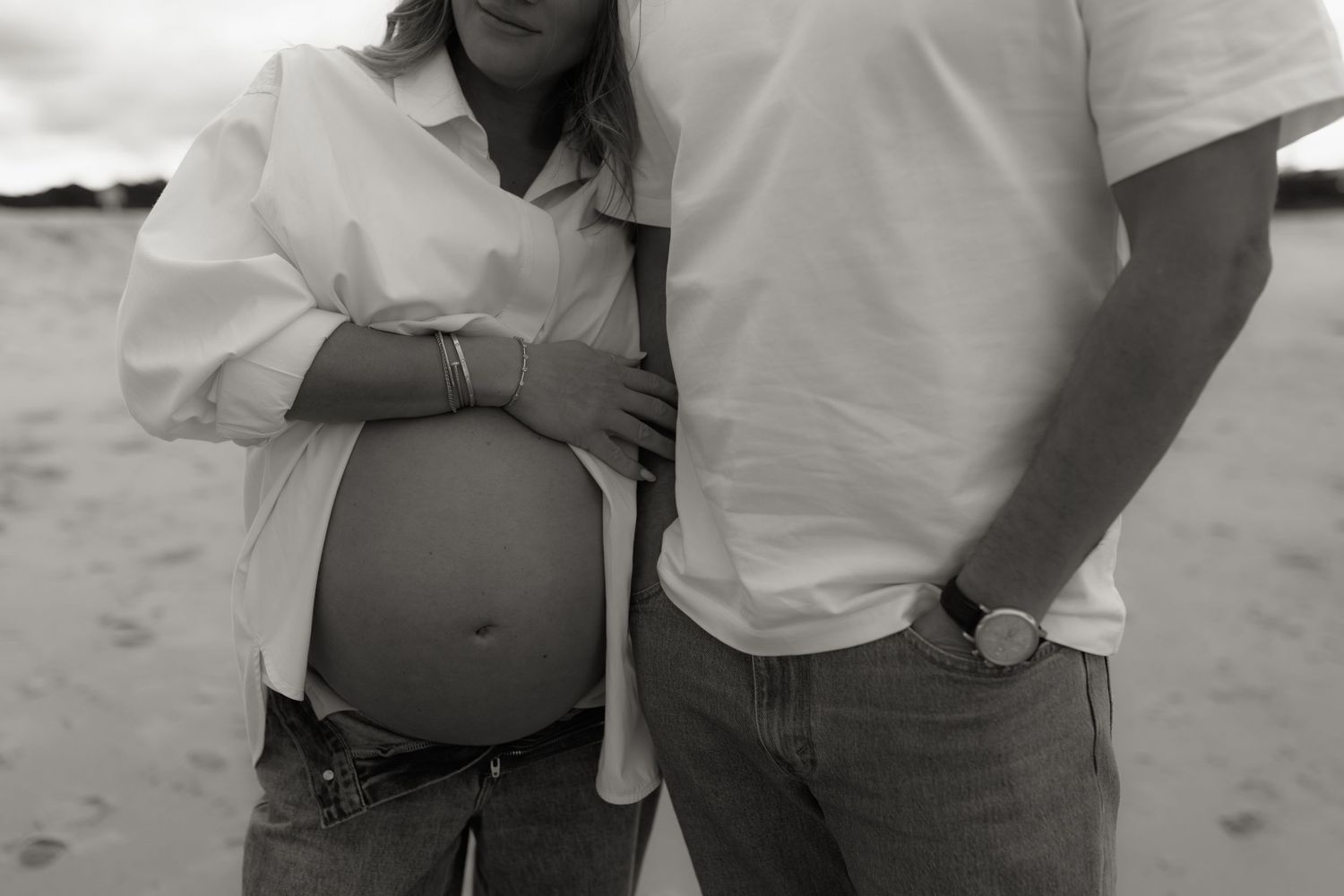 Black and white maternity photo of a pregnant woman and partner standing on a beach, showcasing her baby bump.
