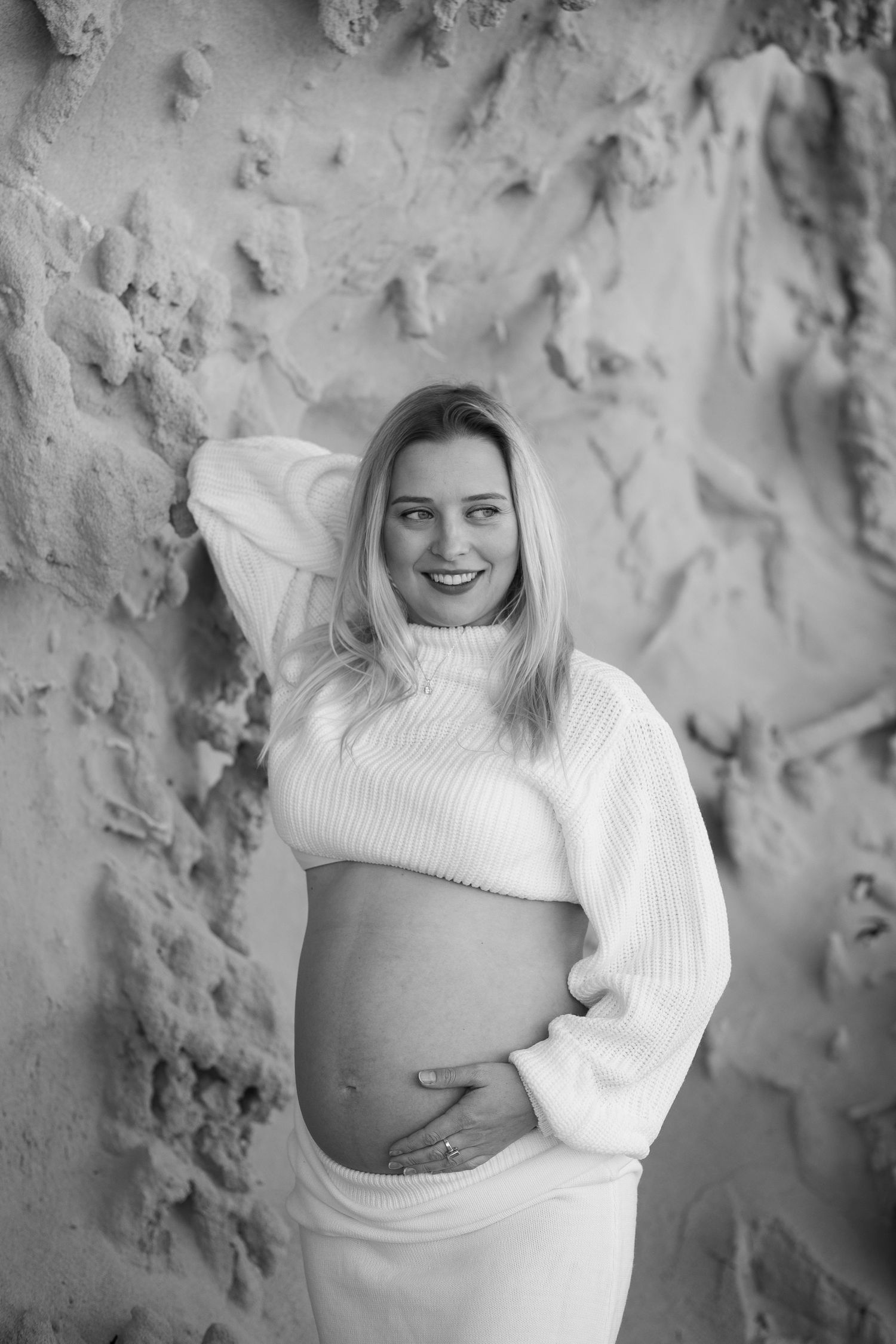 Maternity photo with person wearing white sweater against textured rock wall.