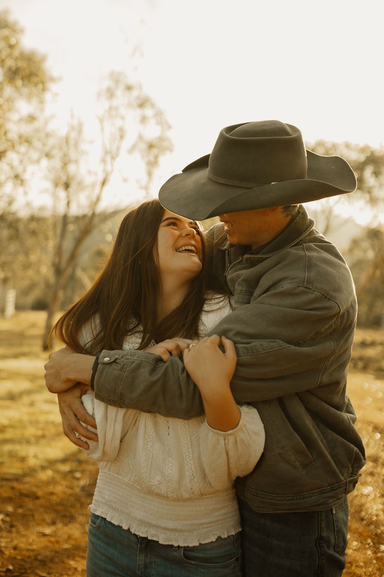 A couple shares a joyful embrace outdoors at sunset, with one wearing a cowboy hat and denim jacket in a rustic setting.