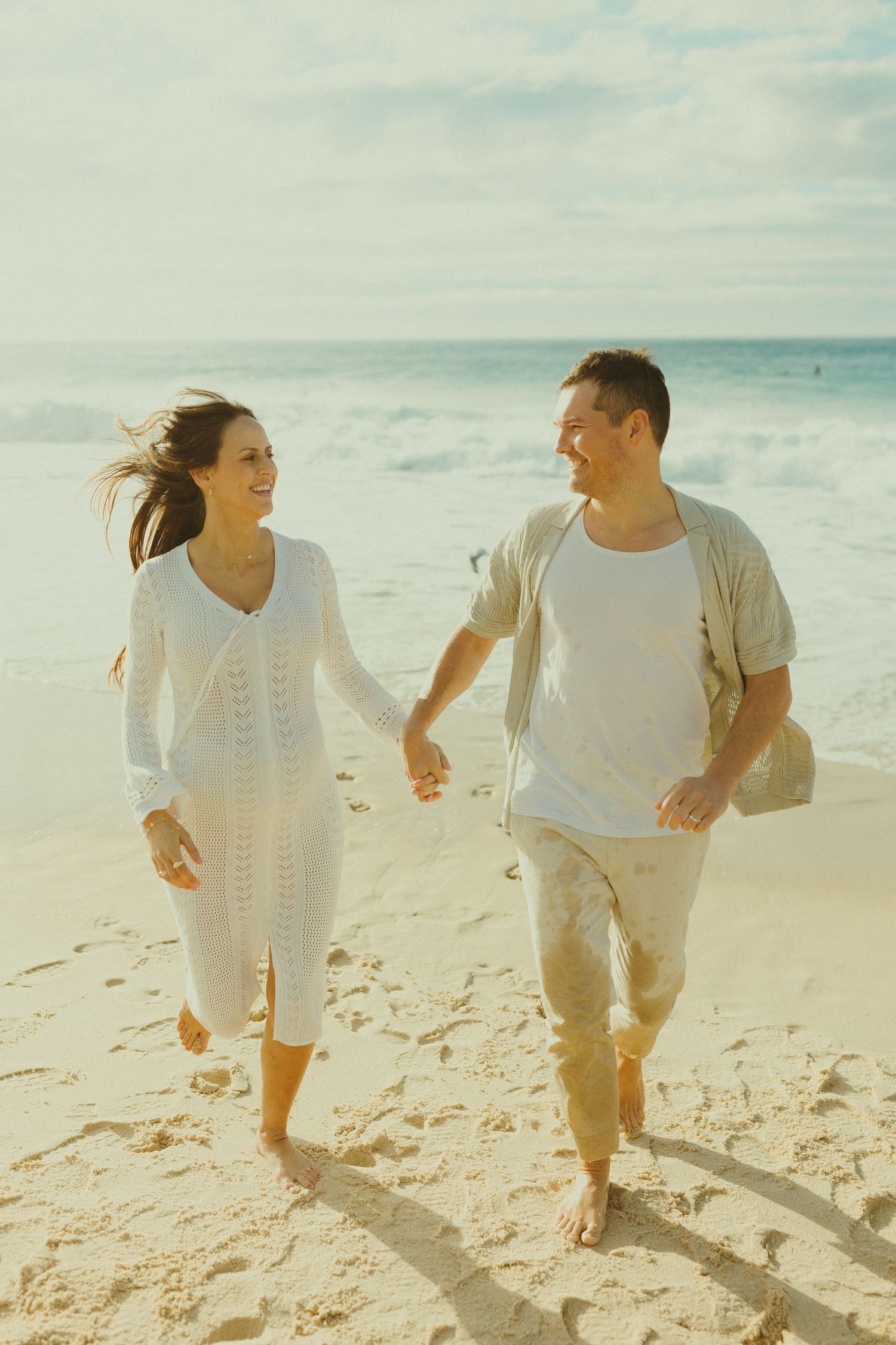 A couple holds hands while walking and laughing together on a sandy beach in matching light-colored outfits.