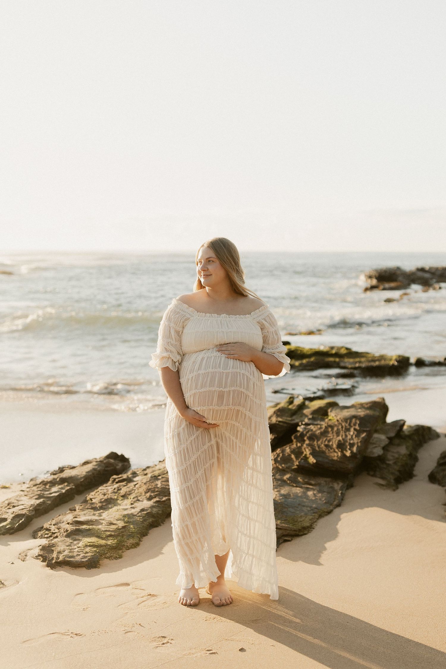Pregnant woman in white lace outfit standing on sandy beach near rocks, cradling her baby bump at sunset.