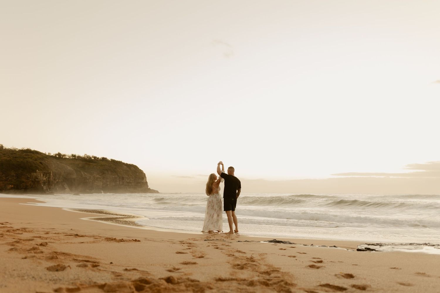 Couple holding hands at sunset on a sandy beach with ocean waves and rocky headland in the background.