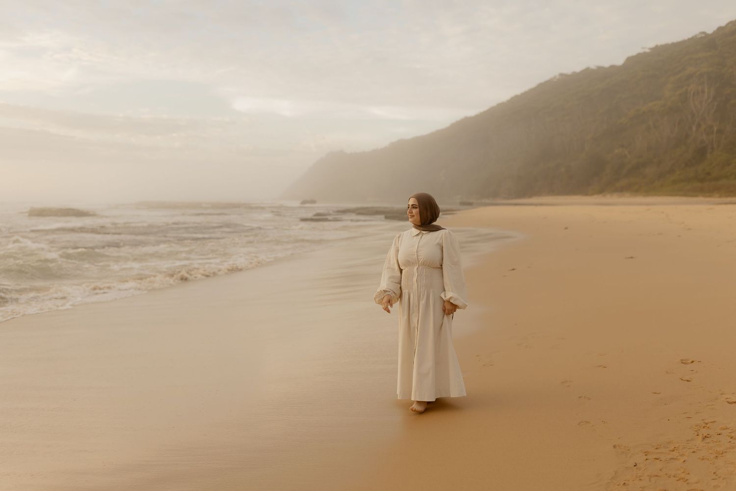 Woman in white dress walking on a sandy beach with misty mountains and ocean waves in the background.