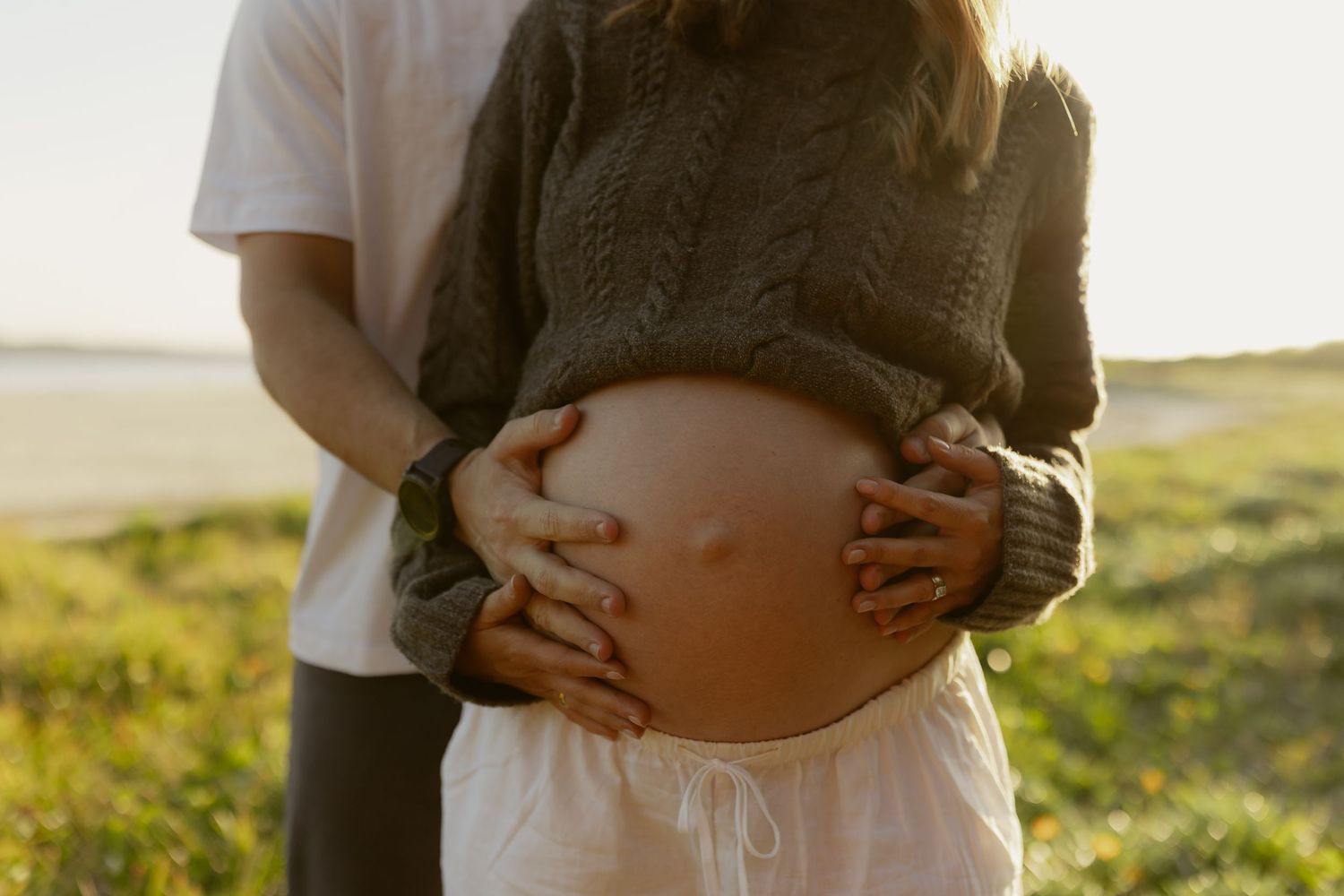 Close-up of a pregnant woman's bare belly being cradled by a couple's hands in golden sunlight.