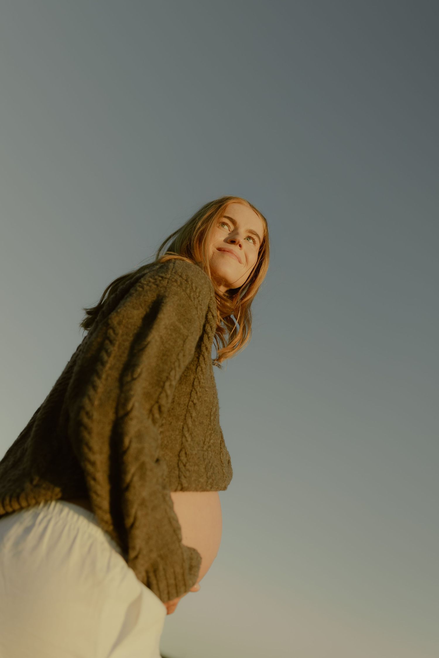 Pregnant woman in olive knit sweater and white skirt smiling outdoors at golden hour, low angle shot against clear sky.