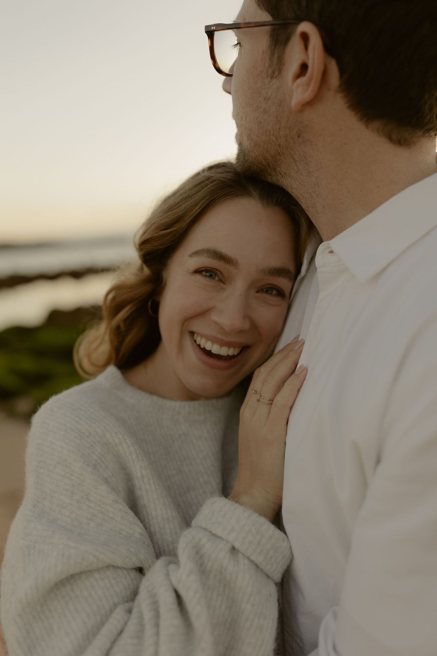 Laughing woman in grey sweater embraces man in white shirt during golden hour beach photoshoot.