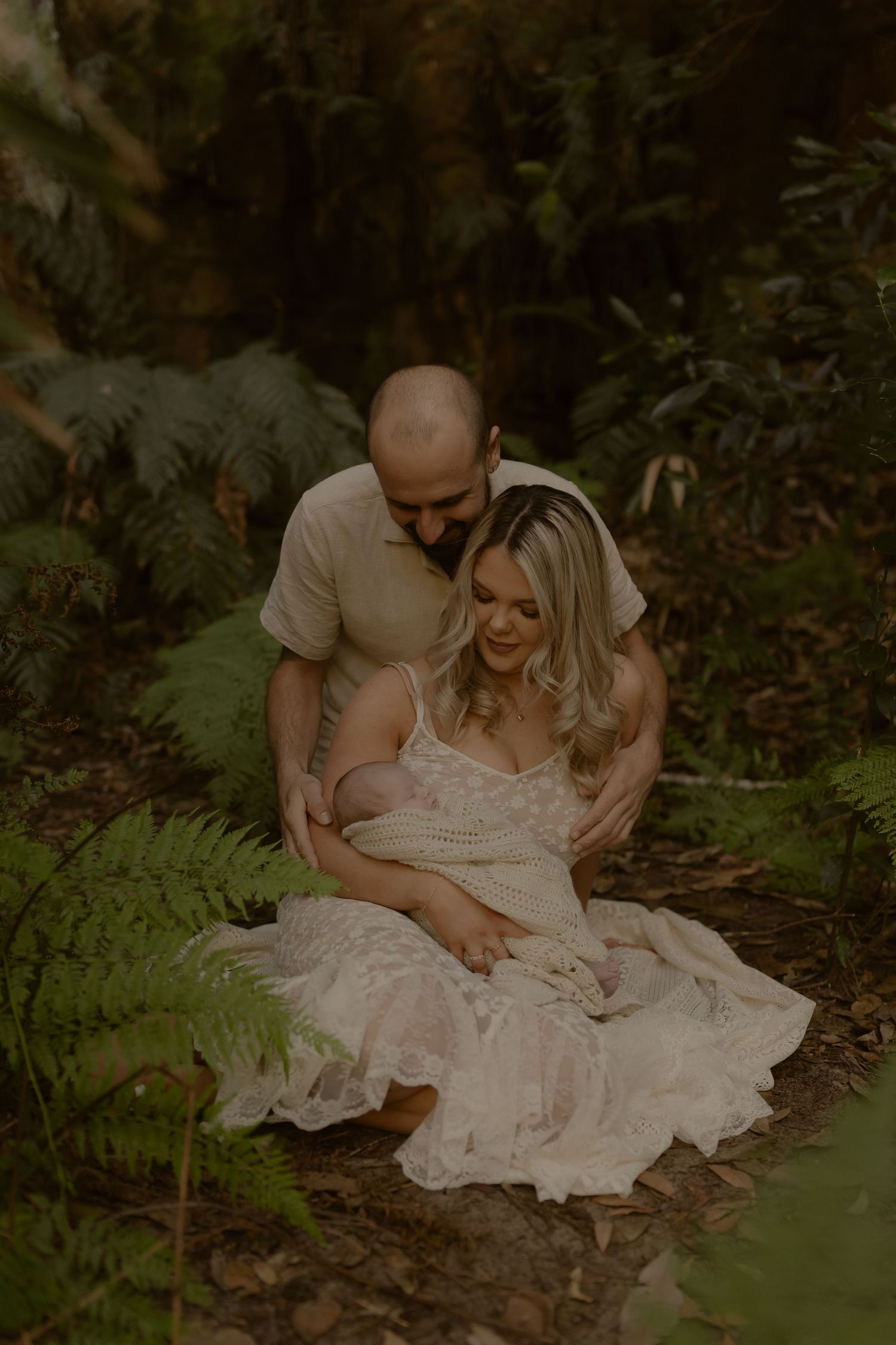 Couple shares intimate moment in lush fern forest, father embracing mother from behind as she cradles newborn.