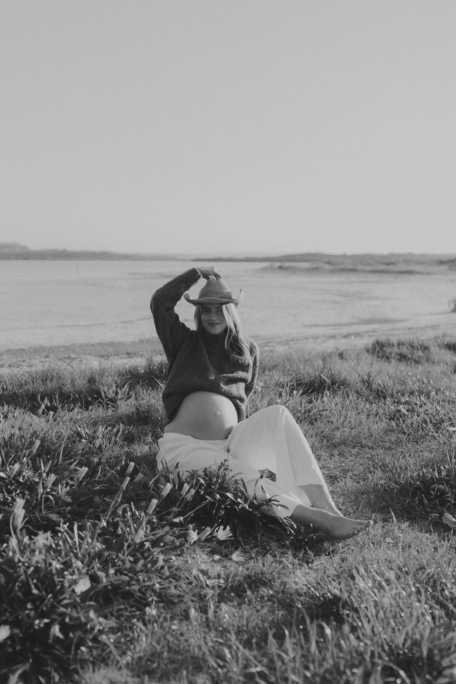 Black and white photo of a pregnant woman sitting on grass near water, wearing a cowboy hat and flowing skirt.
