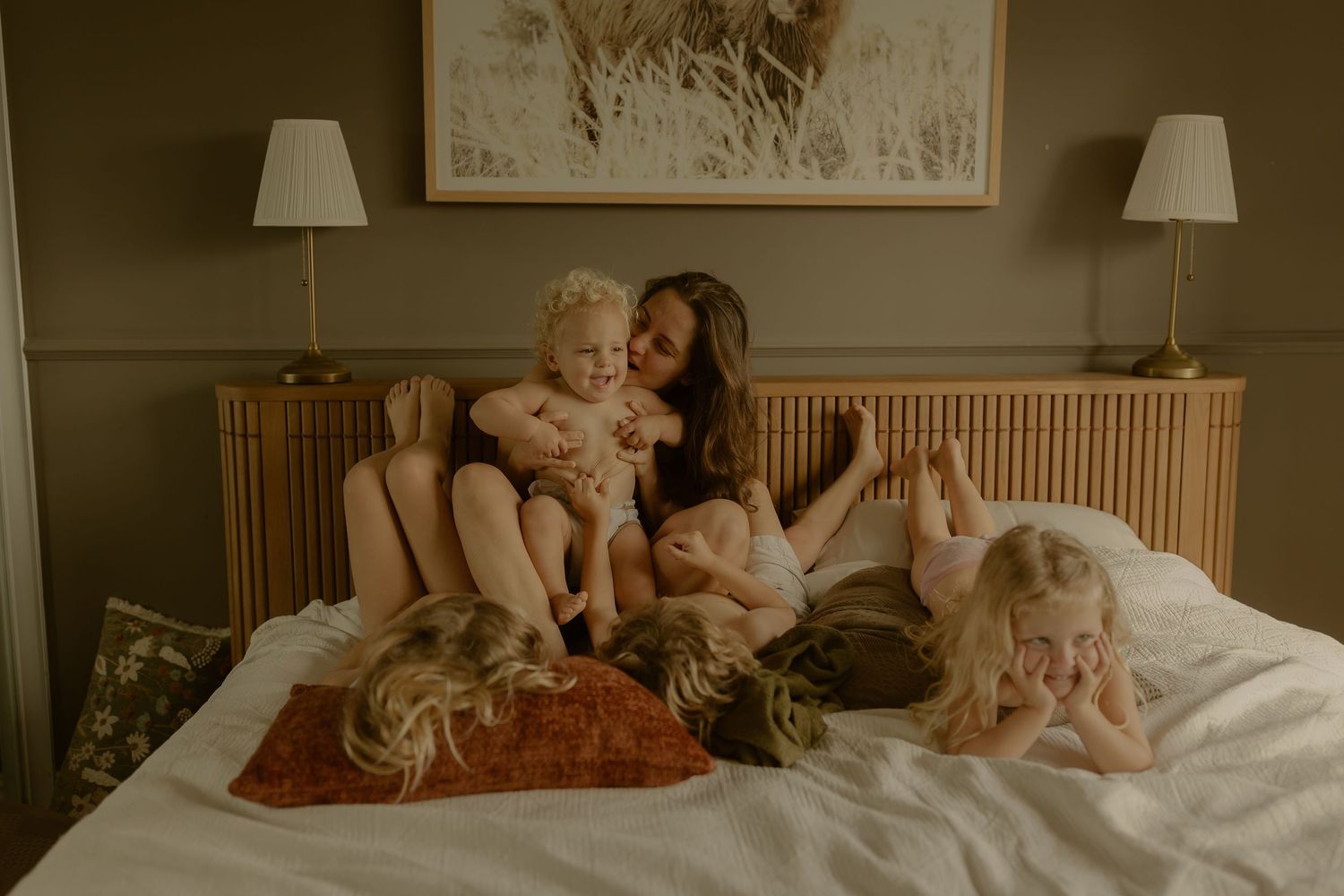 A mother sits on a bed with her three young children, sharing a warm and joyful family moment in a cozy bedroom.