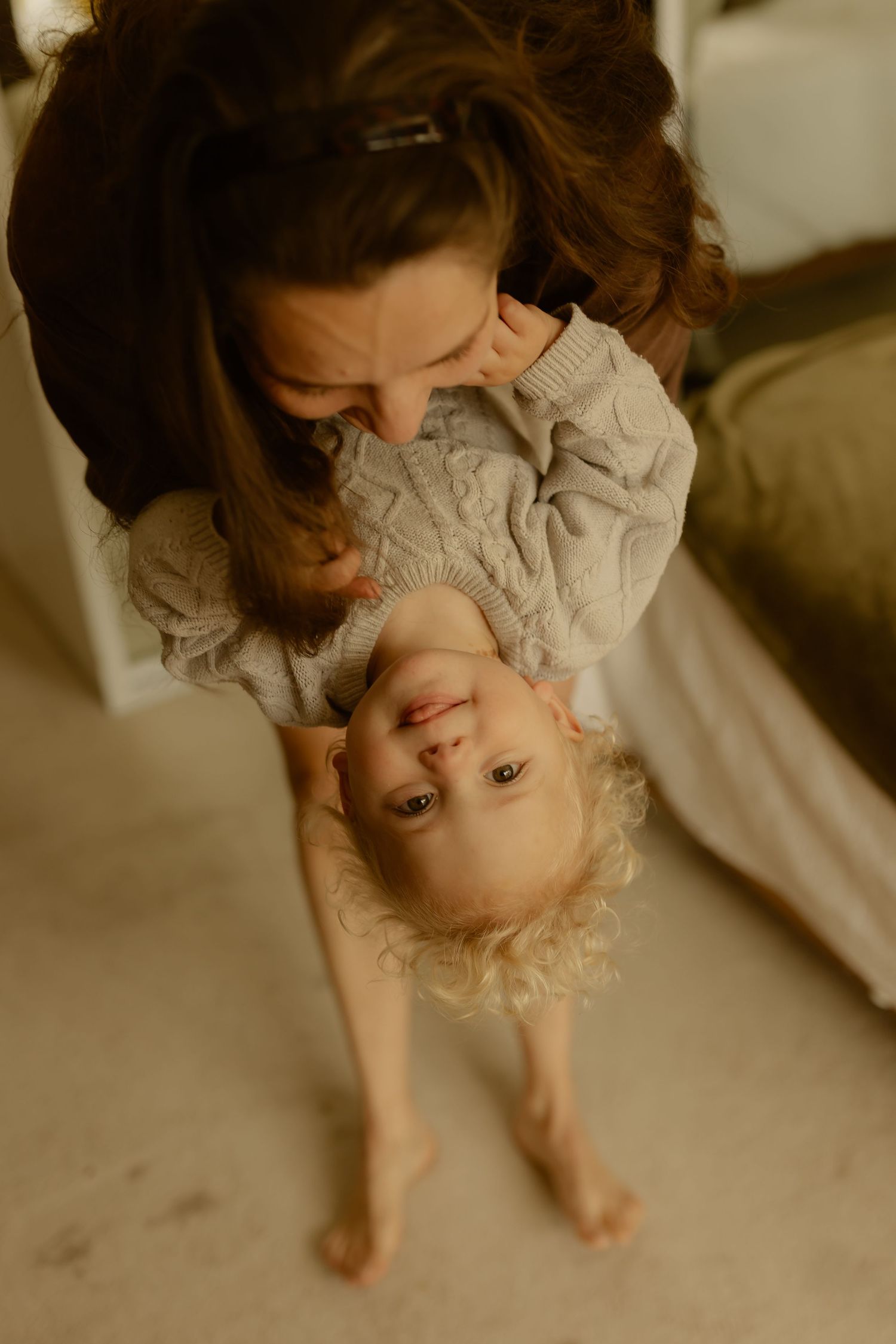 A mother lovingly holds her smiling toddler upside down, both wearing cozy knit sweaters indoors.