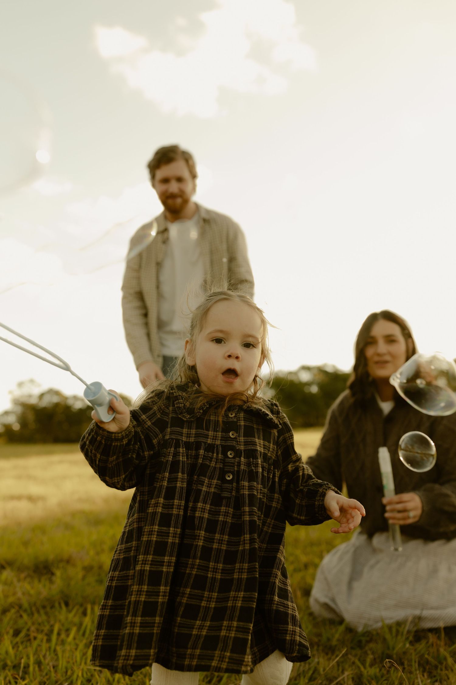 A young girl blows bubbles in a sunny field while her parents watch warmly in the background.