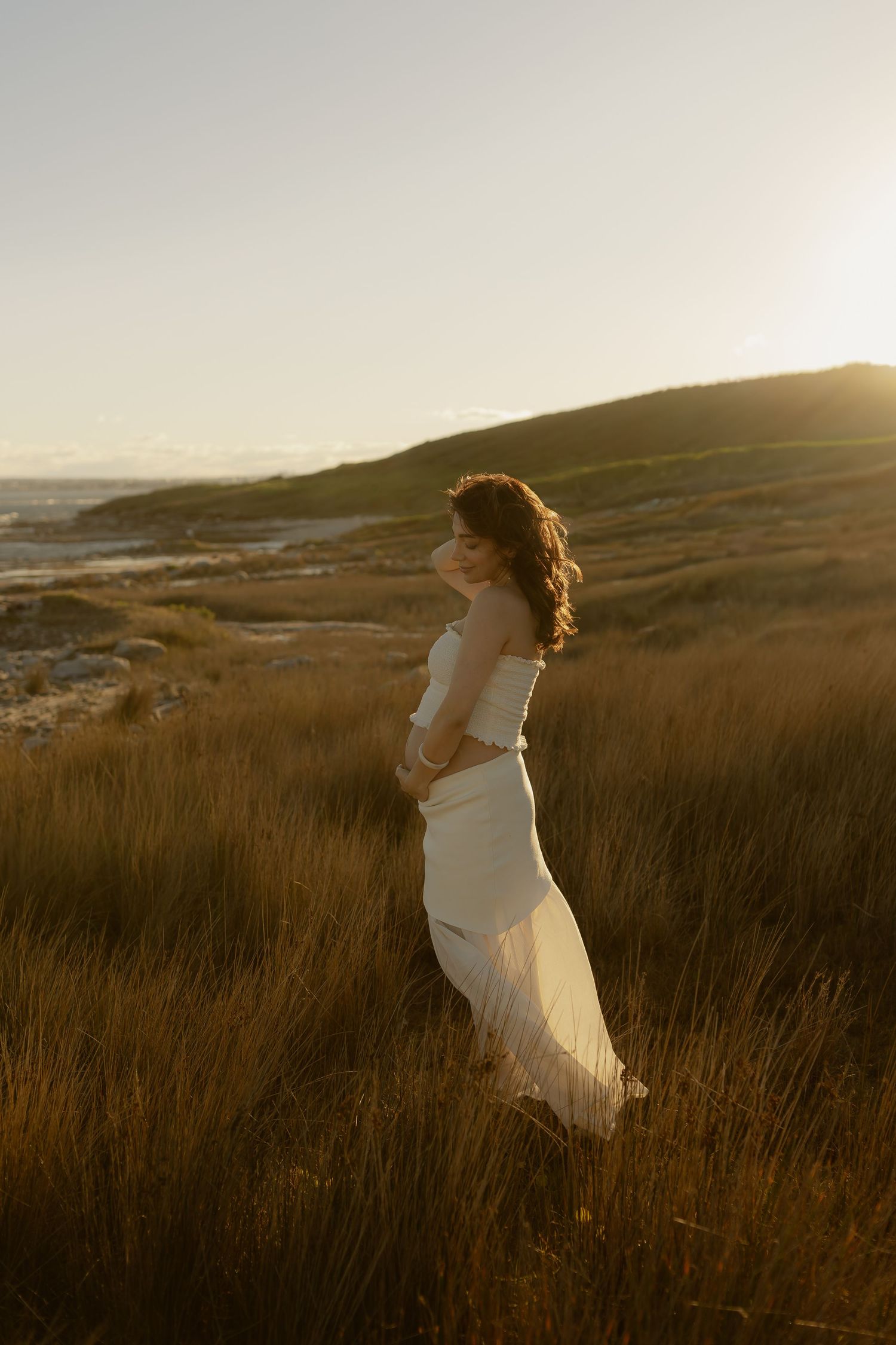 Woman in white dress standing in golden grass fields at sunset with rolling hills and ocean in background.