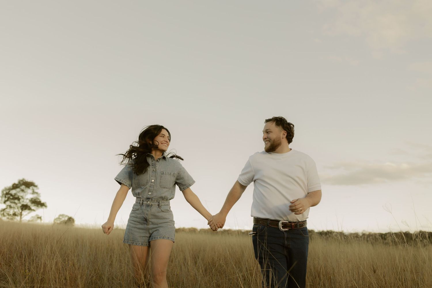 Couple holding hands and smiling at each other while walking through golden grass field at dusk.