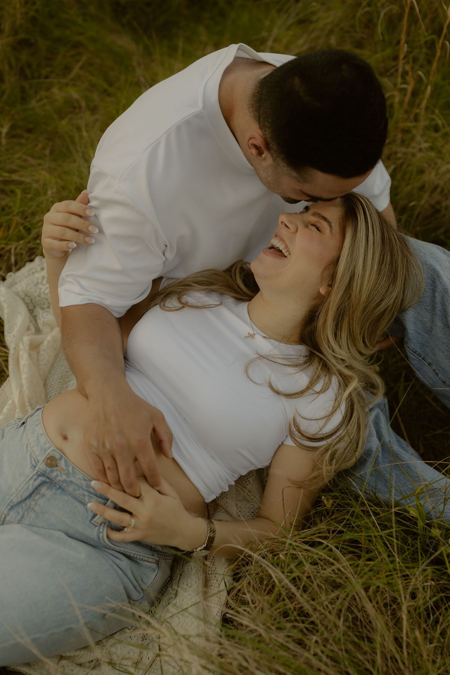 Man kissing laughing pregnant woman as she reclines in tall grass during a golden sunset maternity shoot.