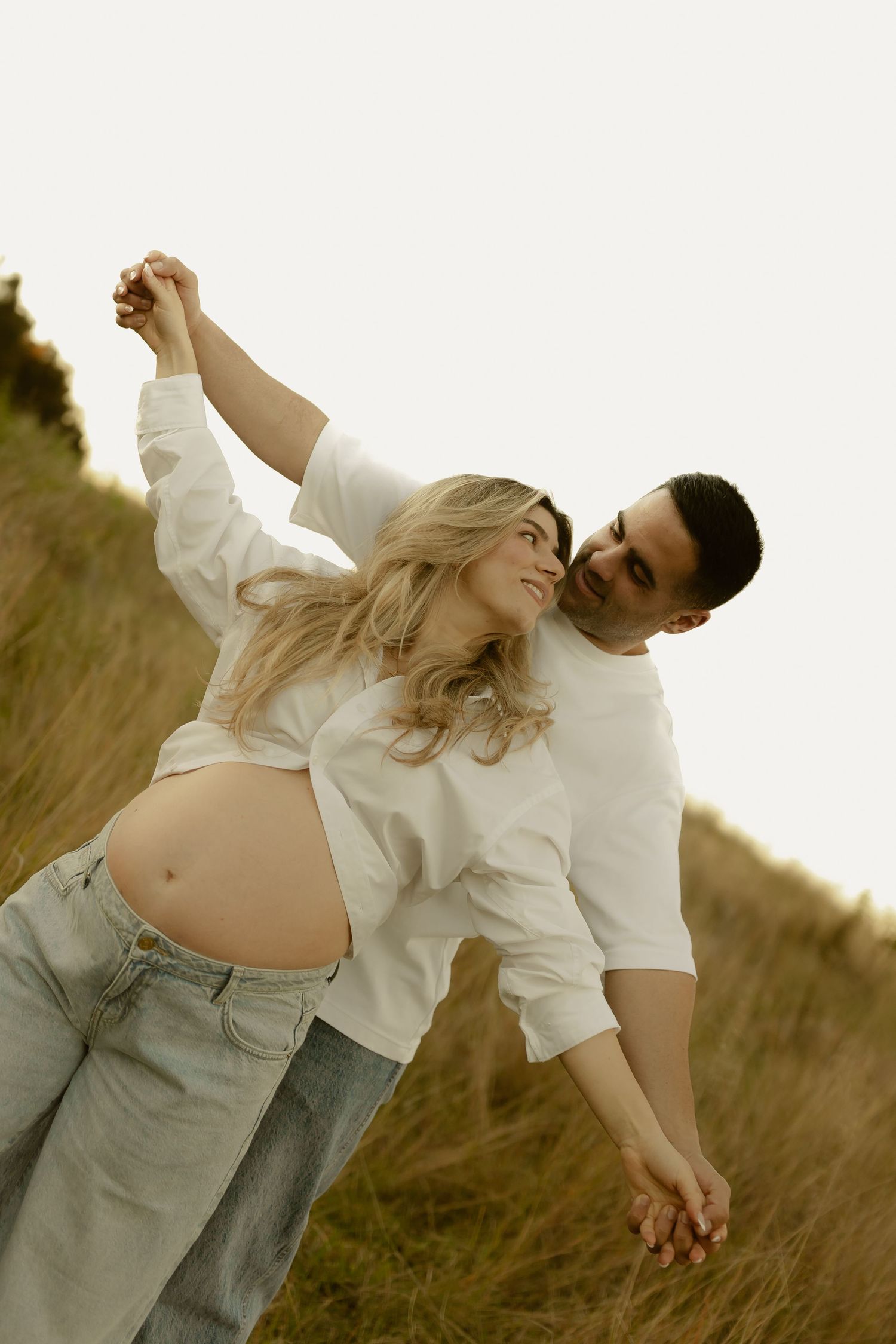 Pregnant couple dancing outdoors in golden grass field, woman in white shirt and jeans with baby bump visible.