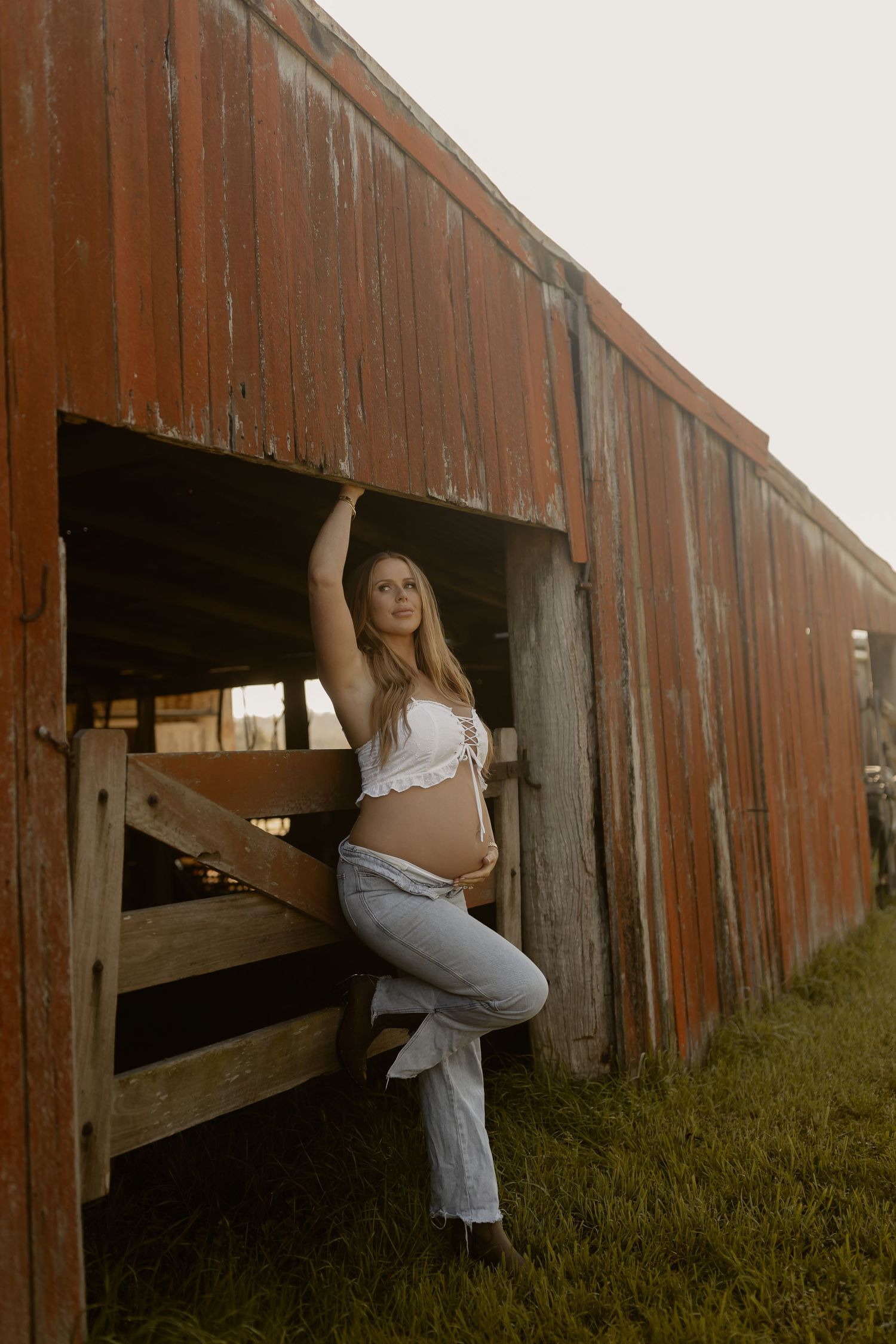 Pregnant woman in white bralette and jeans leans against weathered red barn wall by wooden fence.