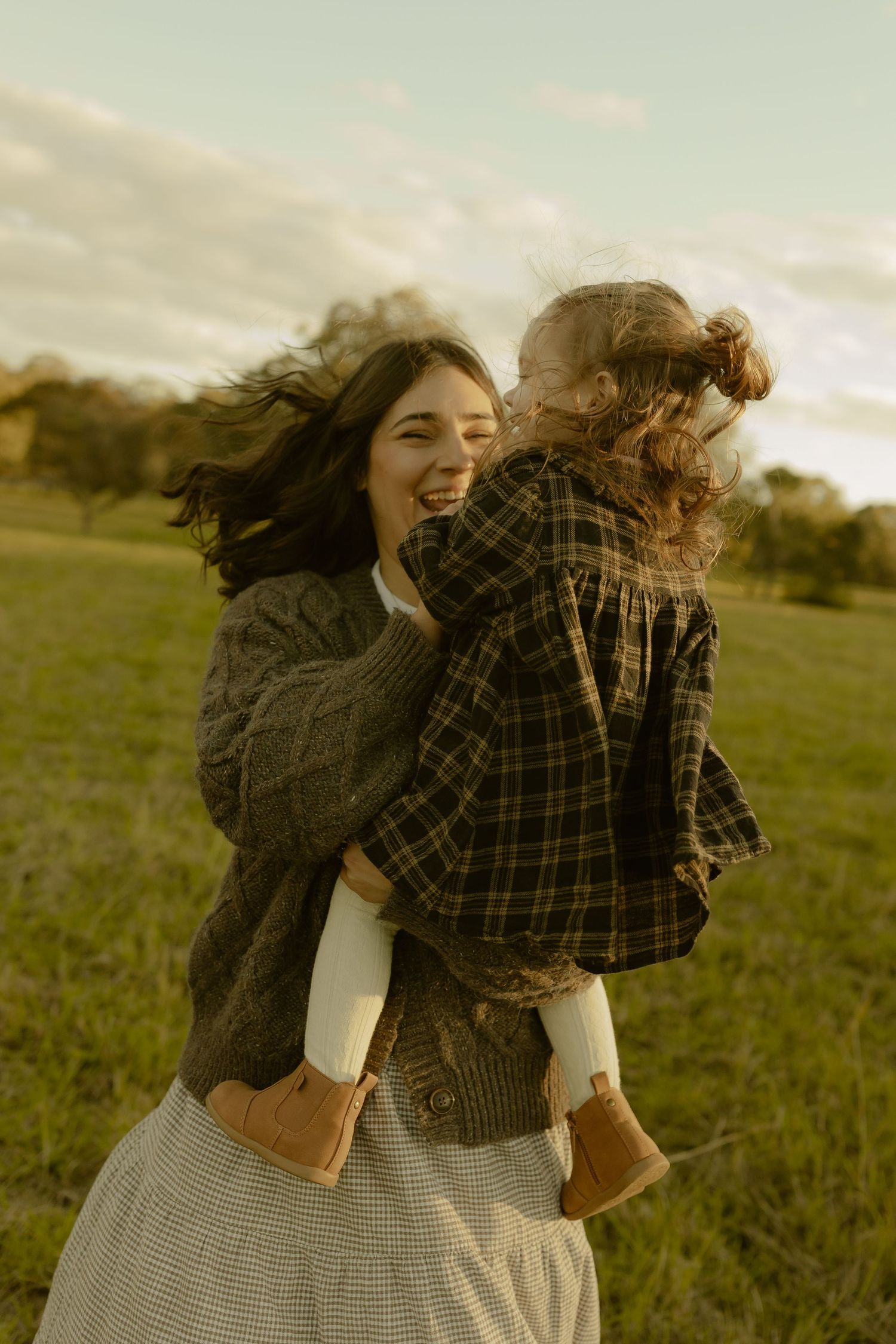 A smiling mother holds her toddler daughter in a sunlit green field, both wearing cozy autumn outfits.