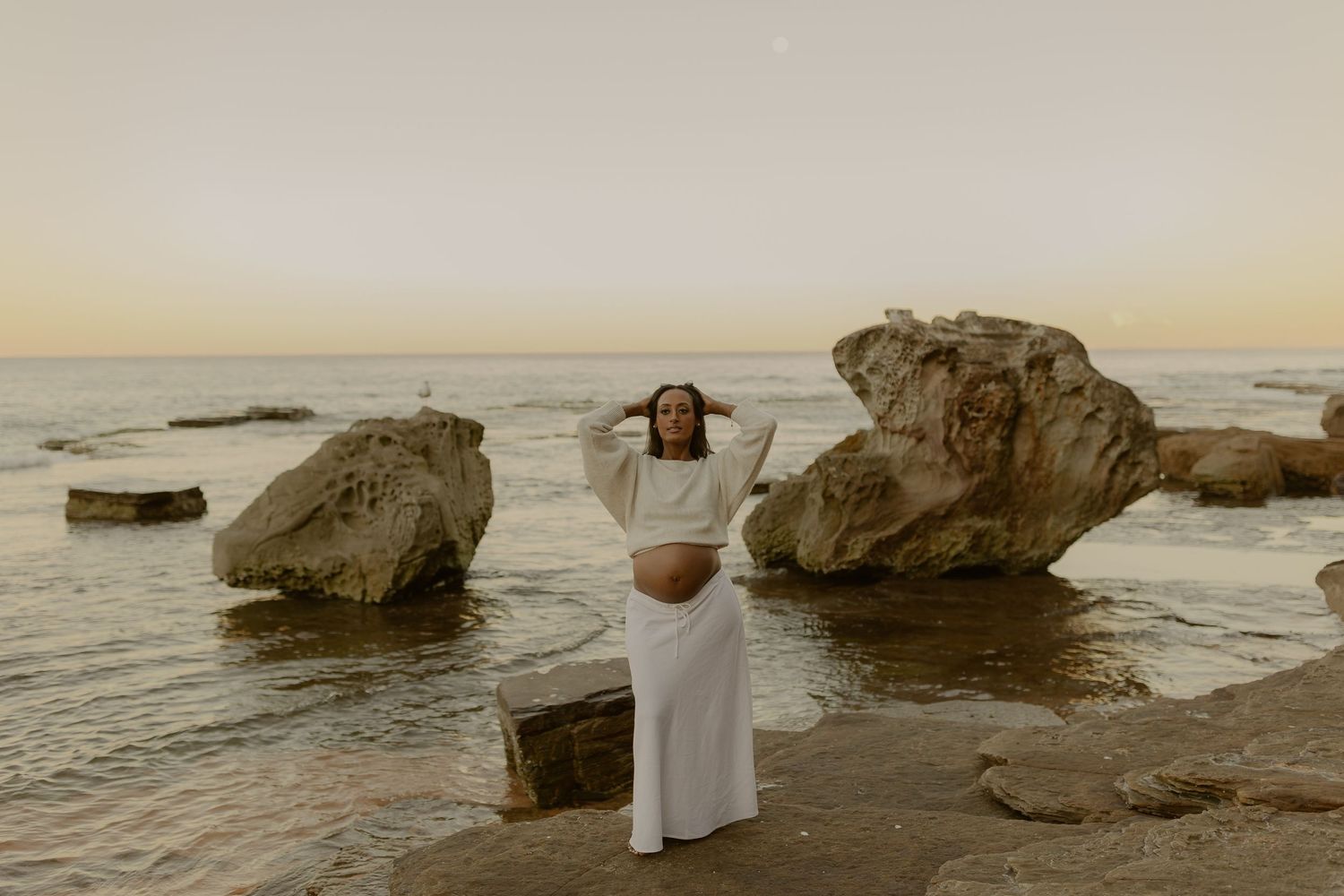 Pregnant woman in white crop top and skirt poses on rocky beach at sunset with large boulders and calm ocean.