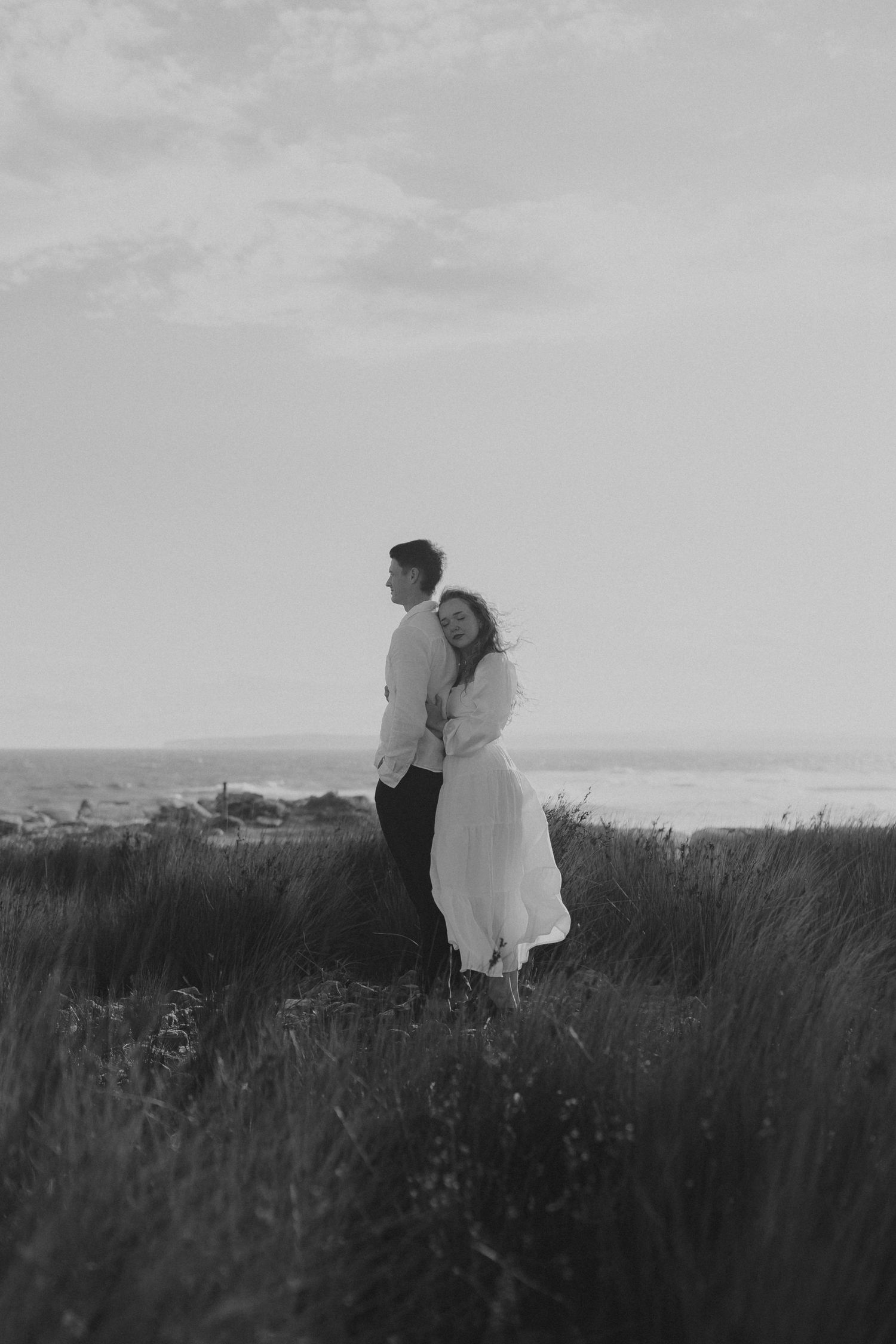 Black and white photo of couple standing together in tall grass with ocean view.