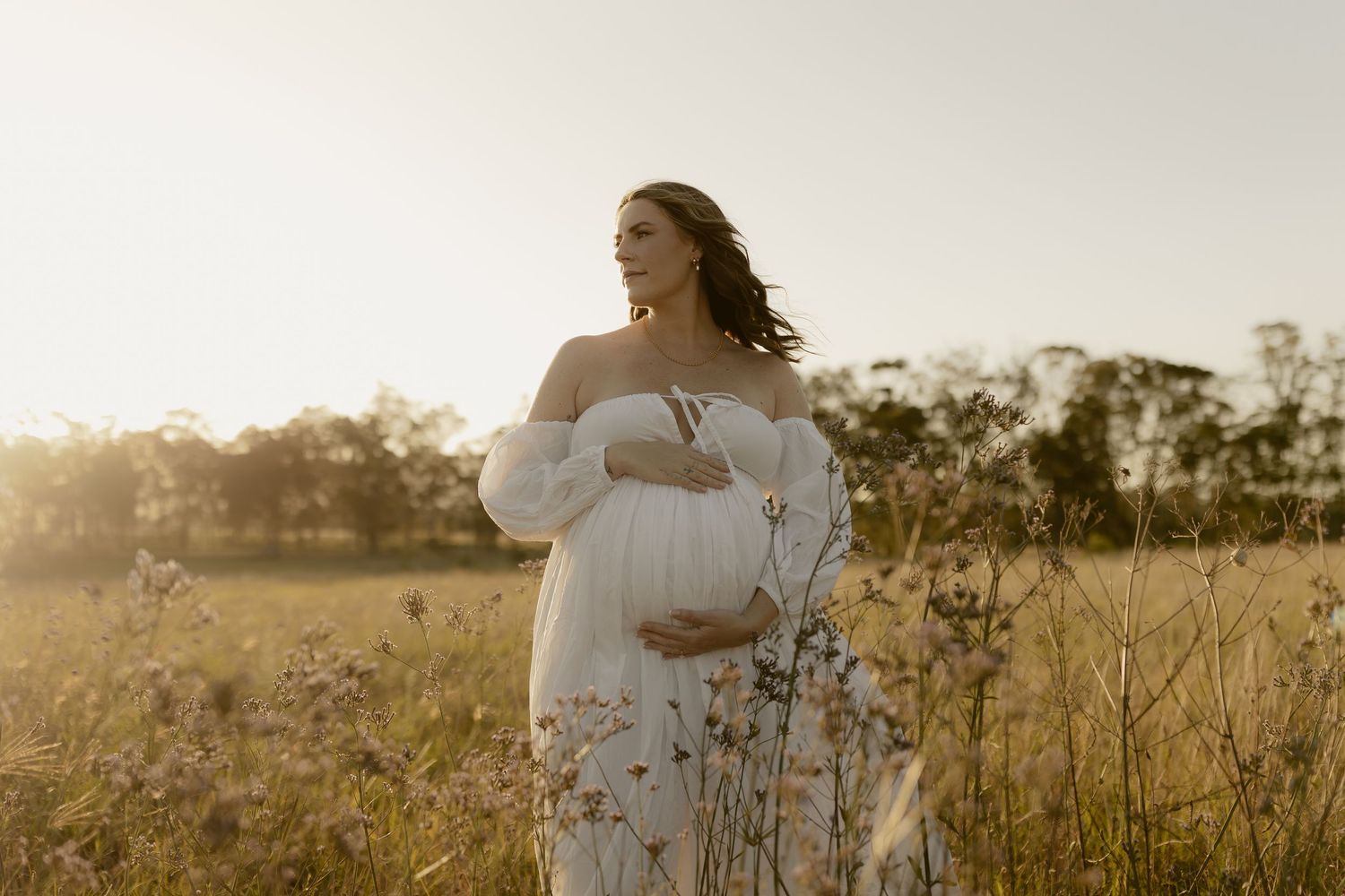 A person in a flowing white dress stands gracefully in a sunlit meadow at golden hour.