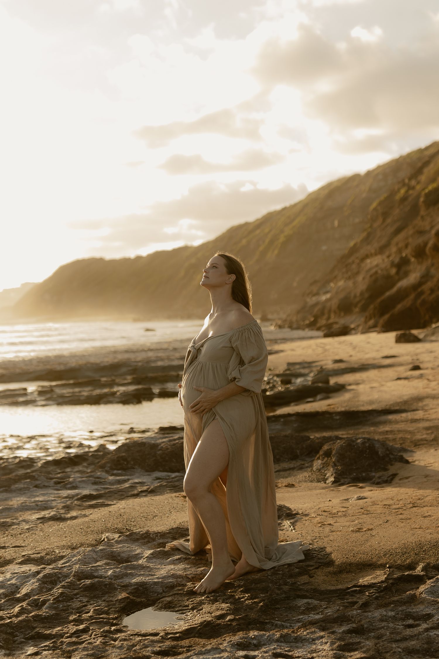 Figure in white dress sits contemplatively on rocky beach at sunset with dramatic coastal cliffs in background.