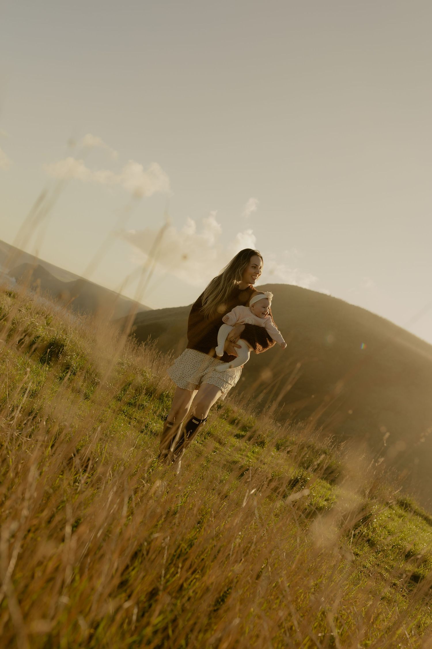 A mother carries her baby through a sunlit golden grass field with rolling green hills in the background.