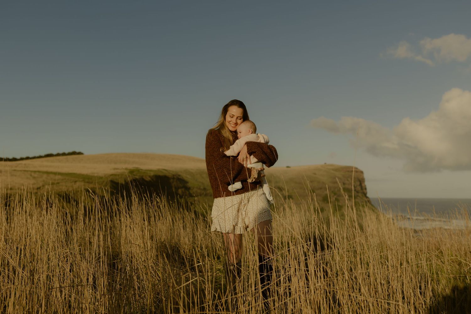 Woman in brown sweater and white skirt holds baby in golden coastal grass field under blue sky.