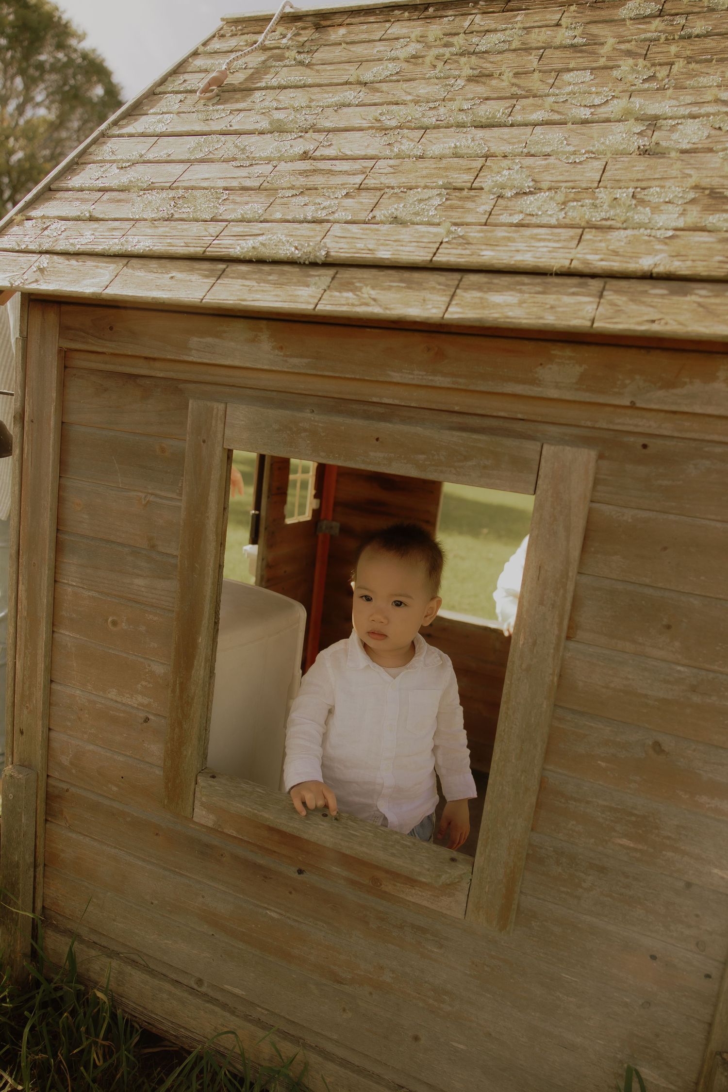 A curious baby peeks through the window of a rustic wooden playhouse outdoors.