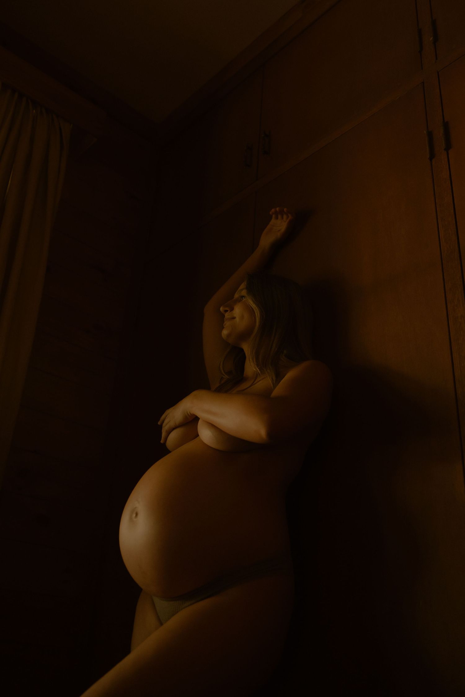 A pregnant woman poses topless in dramatic warm shadow light, arm raised gracefully against a dark wall.
