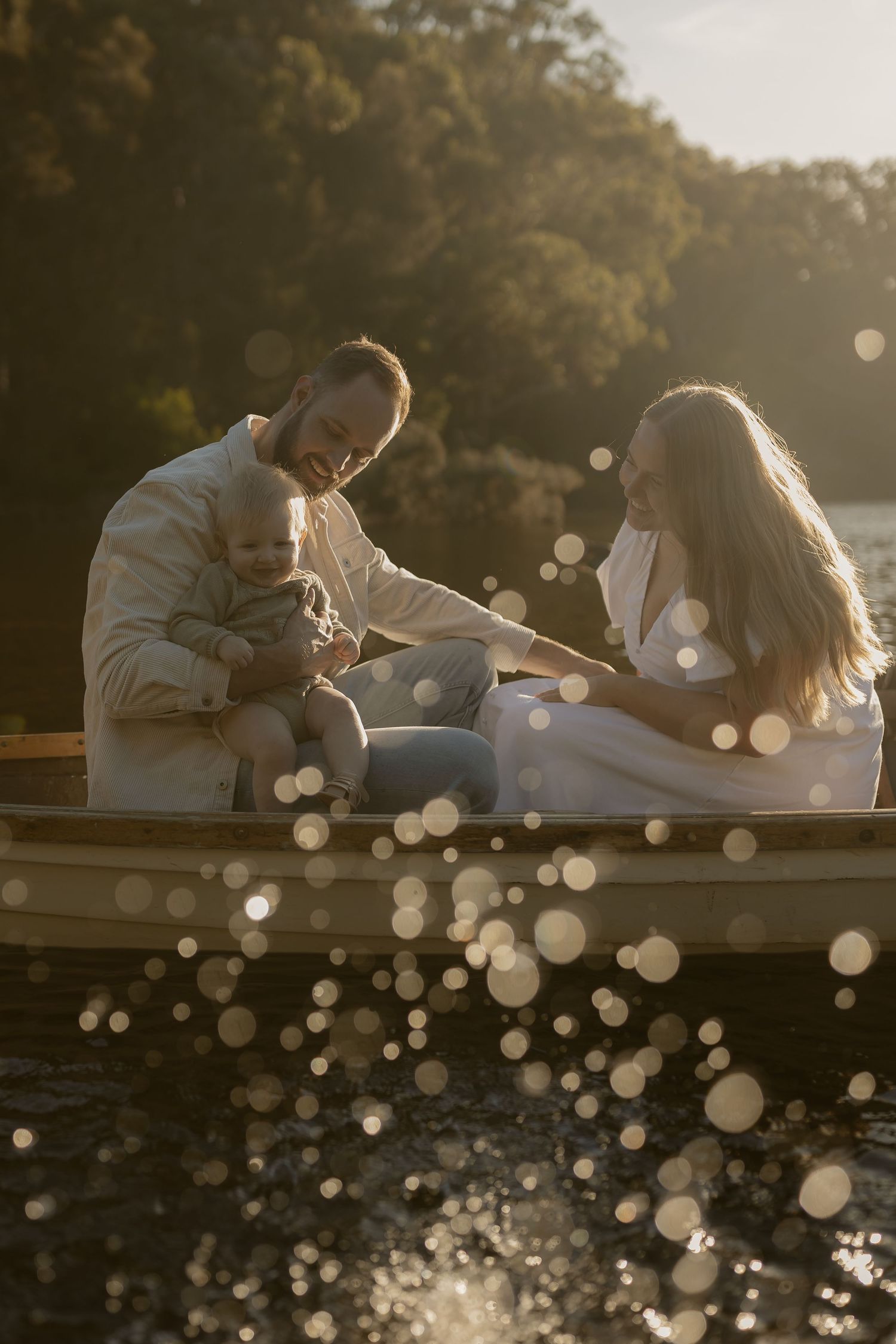 Family with a baby sitting together in a wooden rowboat on a sunlit lake surrounded by bokeh light effects.