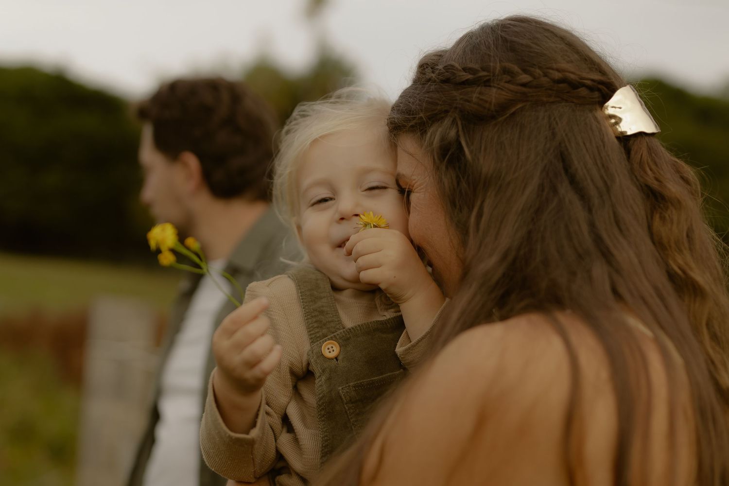 Woman kissing a smiling toddler holding yellow wildflowers outdoors with a man visible in the background.