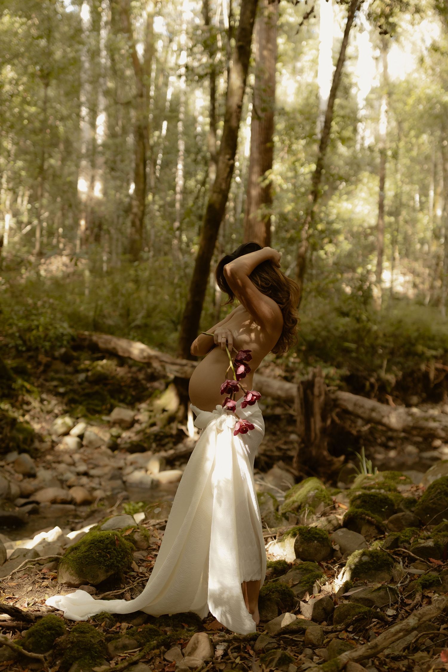 A woman in a flowing white skirt and floral top poses gracefully among mossy rocks in a sunlit forest.
