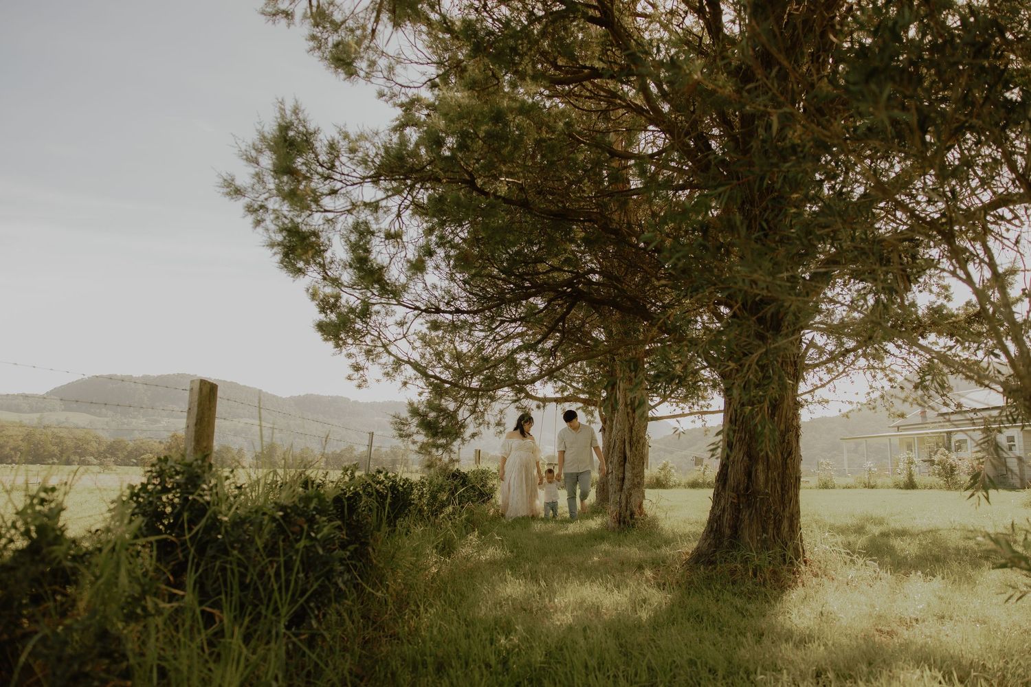 Family of three walking under large trees in a lush green countryside meadow with rolling hills in the background.