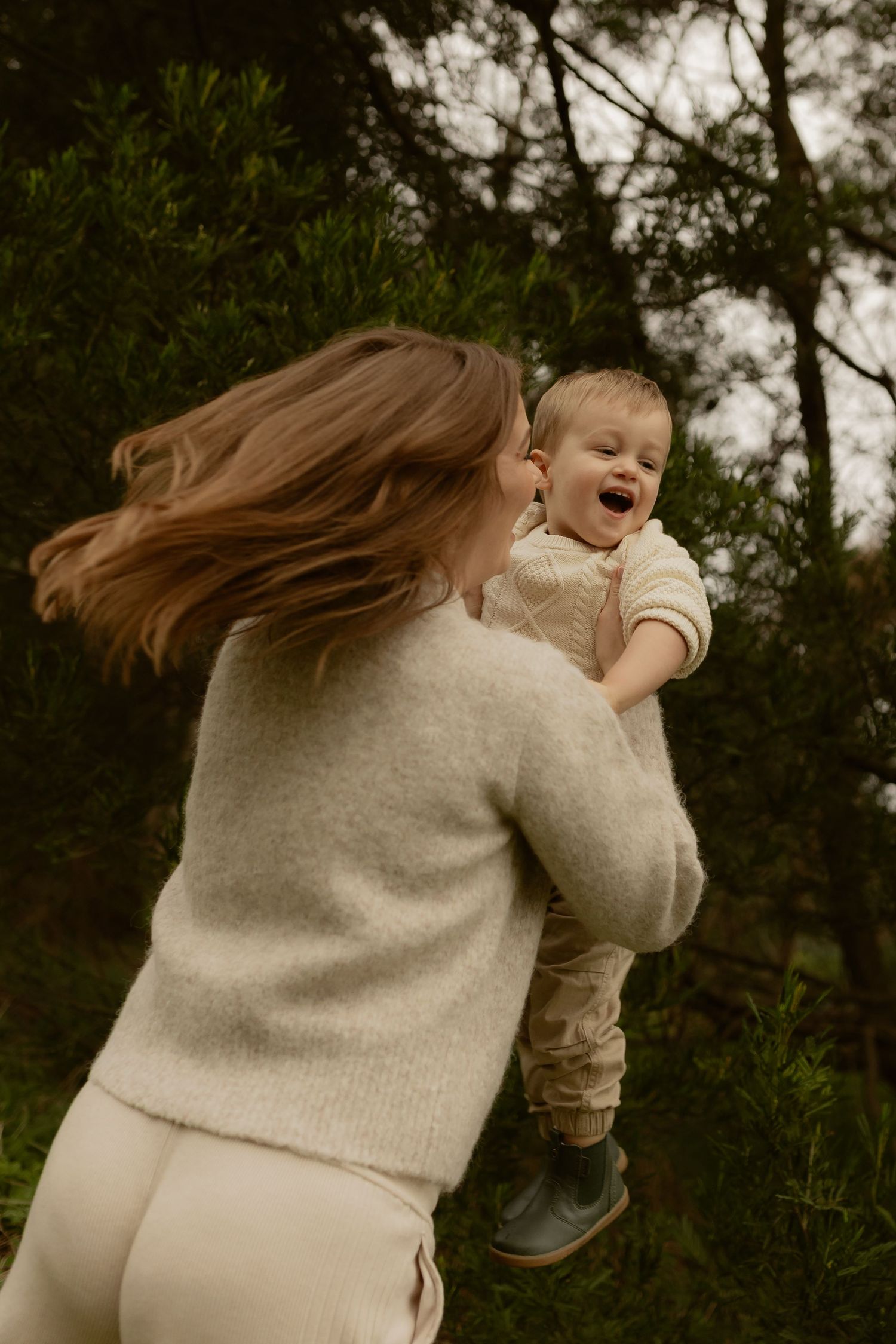 A joyful mother lifts her laughing toddler in the air surrounded by lush green bushes outdoors.