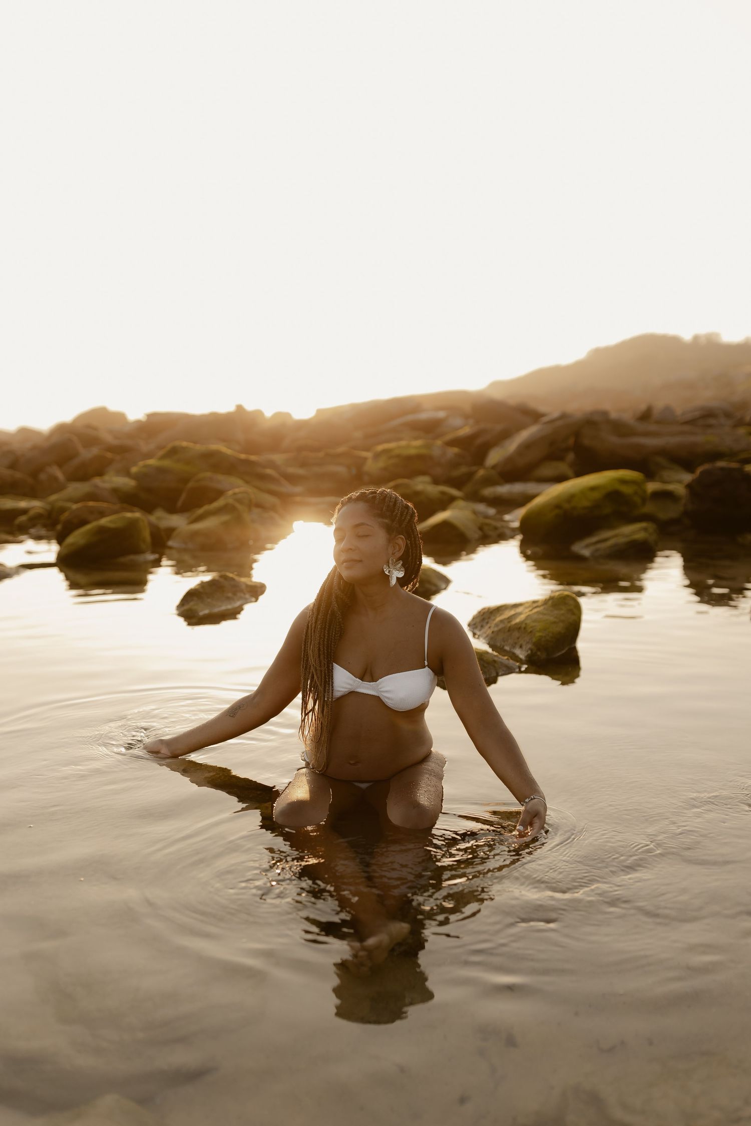 A person in a black swimsuit floats serenely in calm ocean waters at sunset near rocky outcrops.