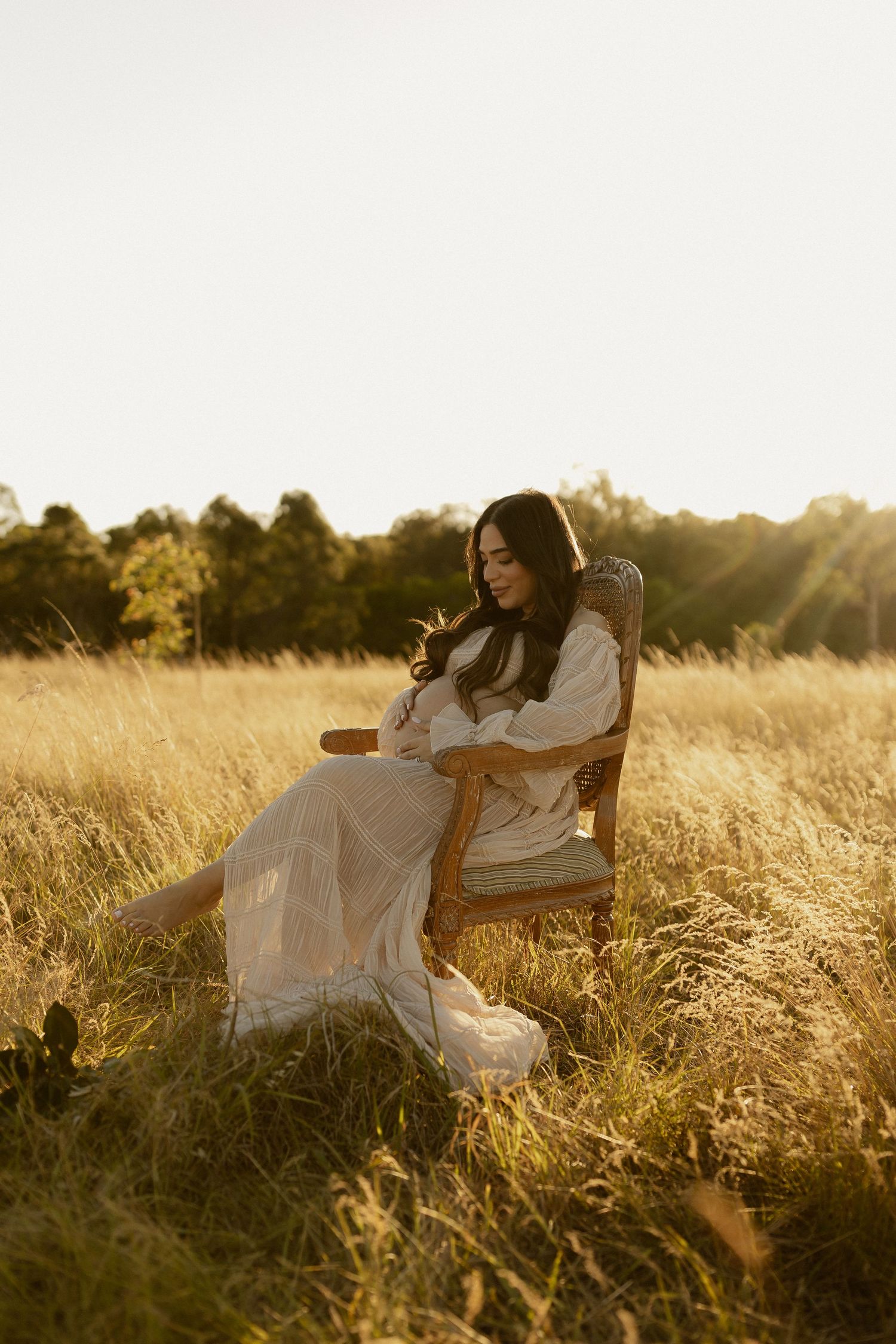 Expectant mother in white dress sitting in vintage chair amid sun-drenched field.