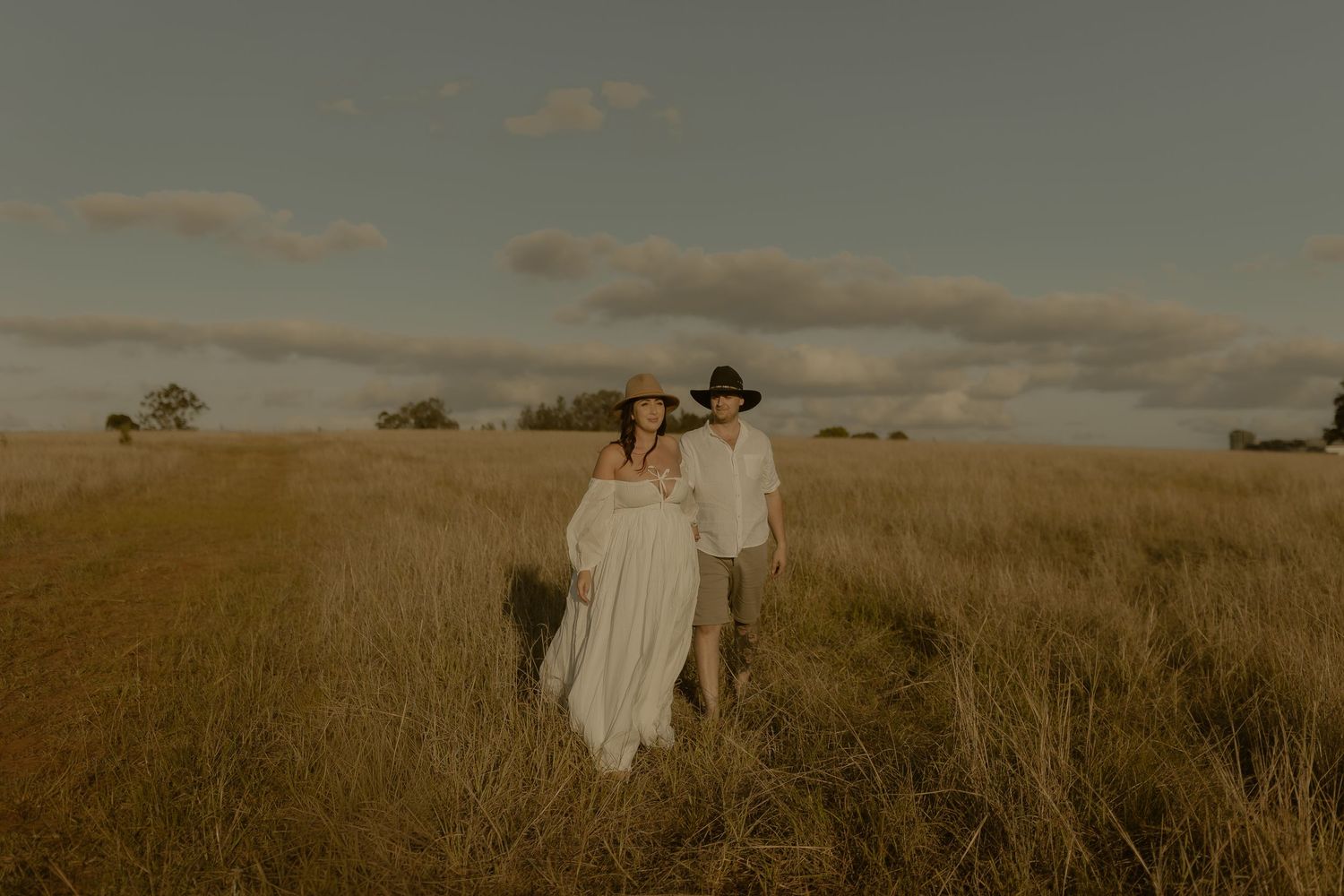 Couple in white outfits walks through golden prairie grass field during sunset with warm lighting.