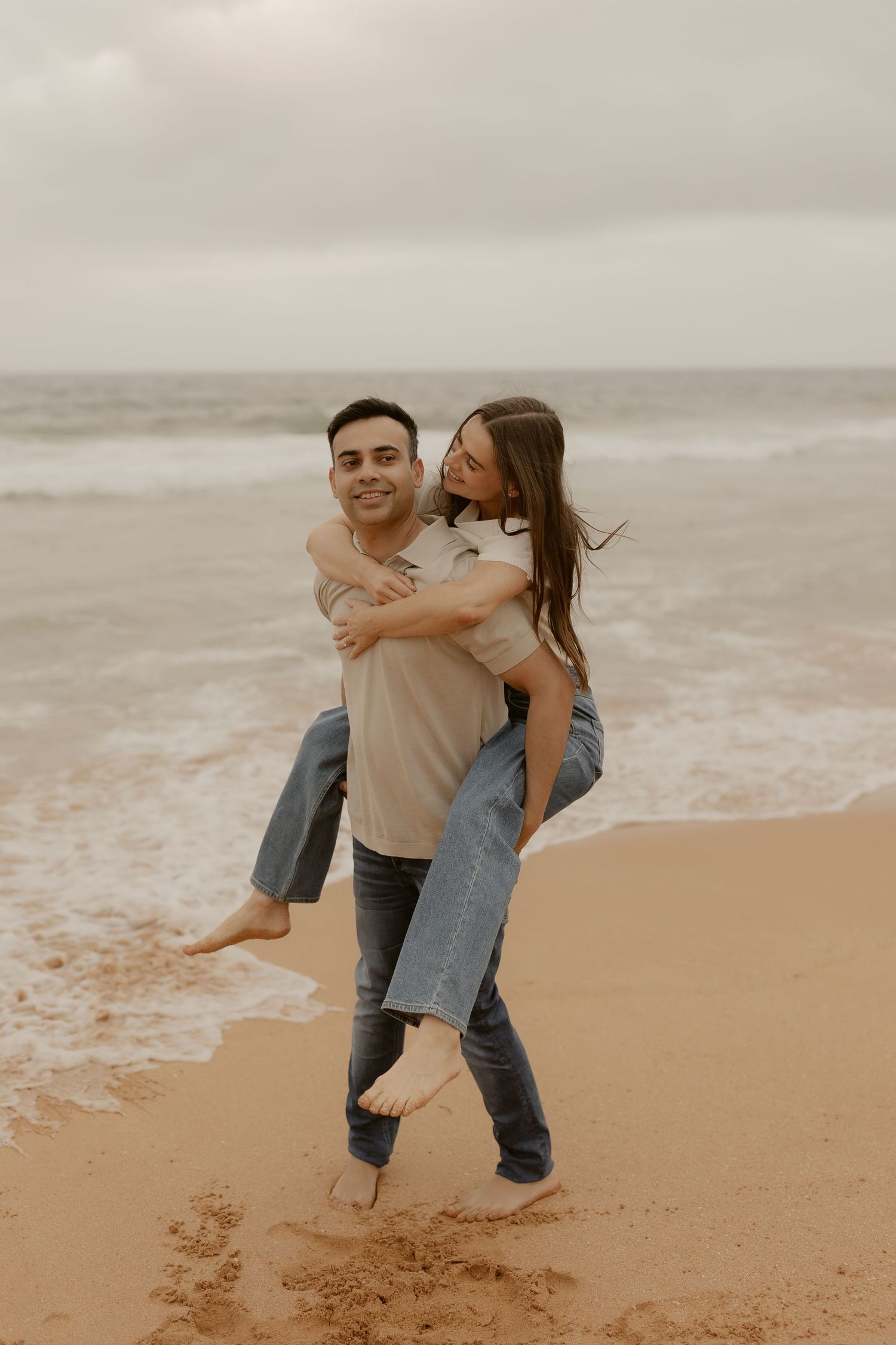 A playful piggyback ride moment on a cloudy beach with waves in the background.