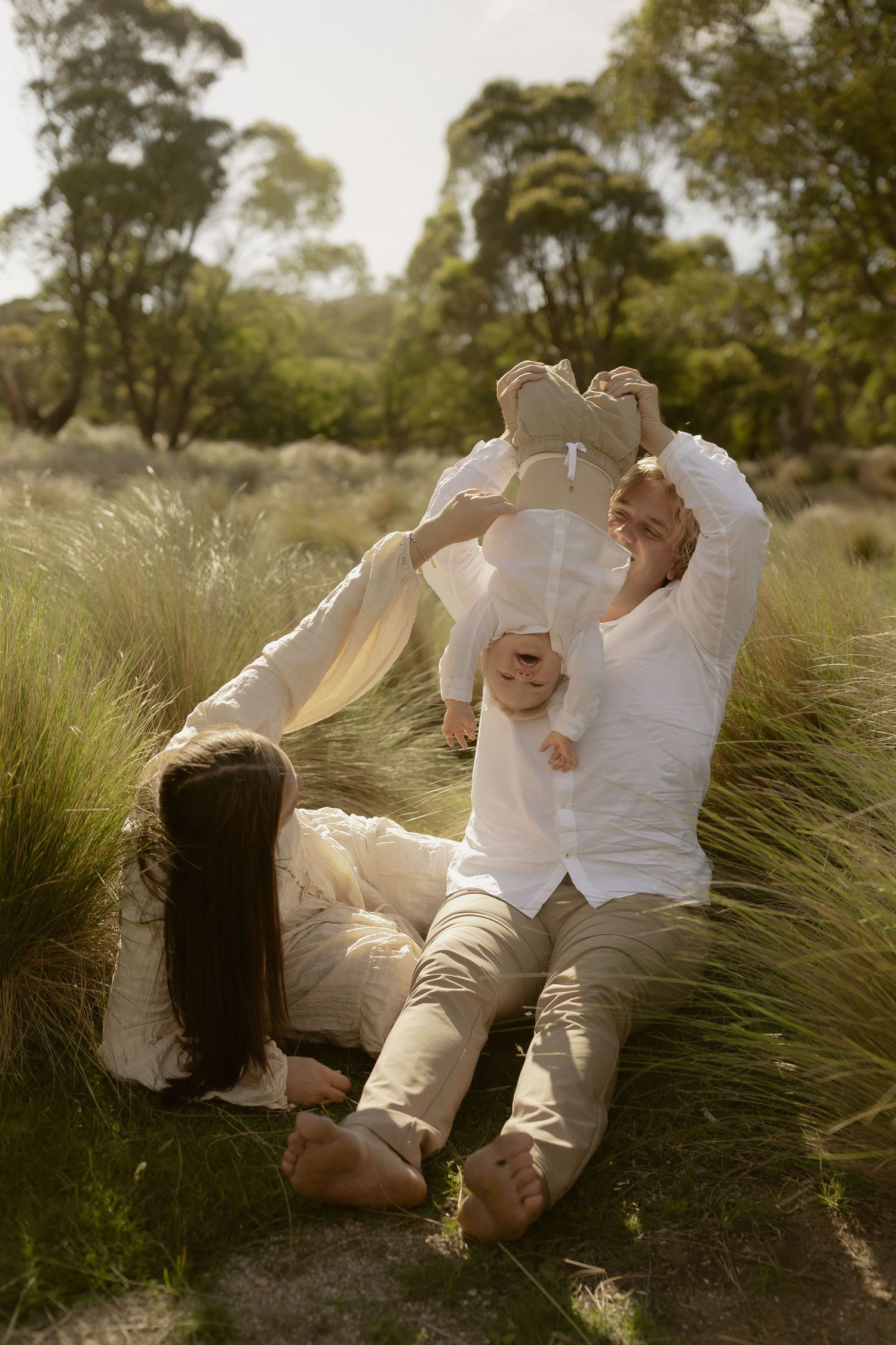 A couple in white clothing lies together in tall grass during a romantic outdoor photoshoot at sunset.