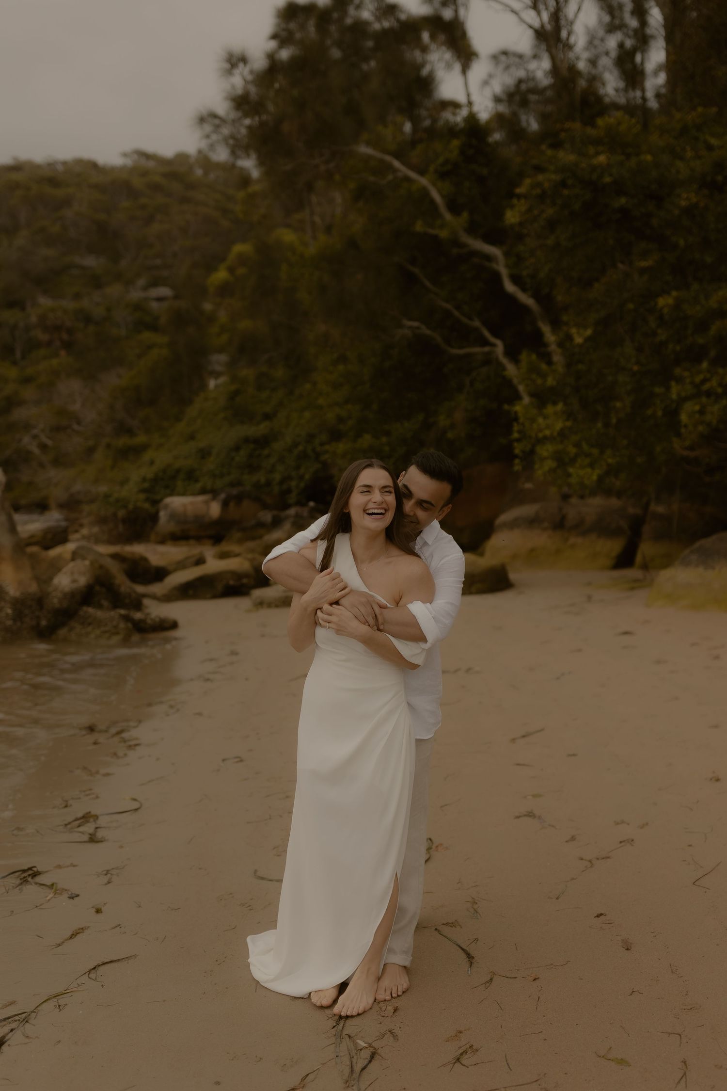 A romantic beach photoshoot featuring a couple embracing on a sandy shore with lush trees in the background.