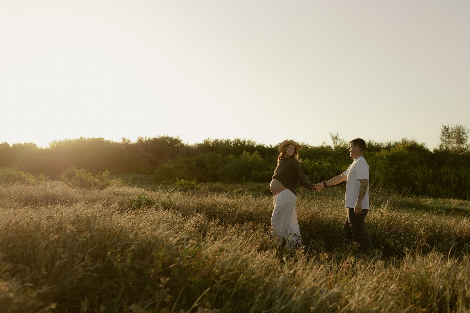 Pregnant couple holding hands in a golden sunlit field during sunset, surrounded by tall grass and green trees.