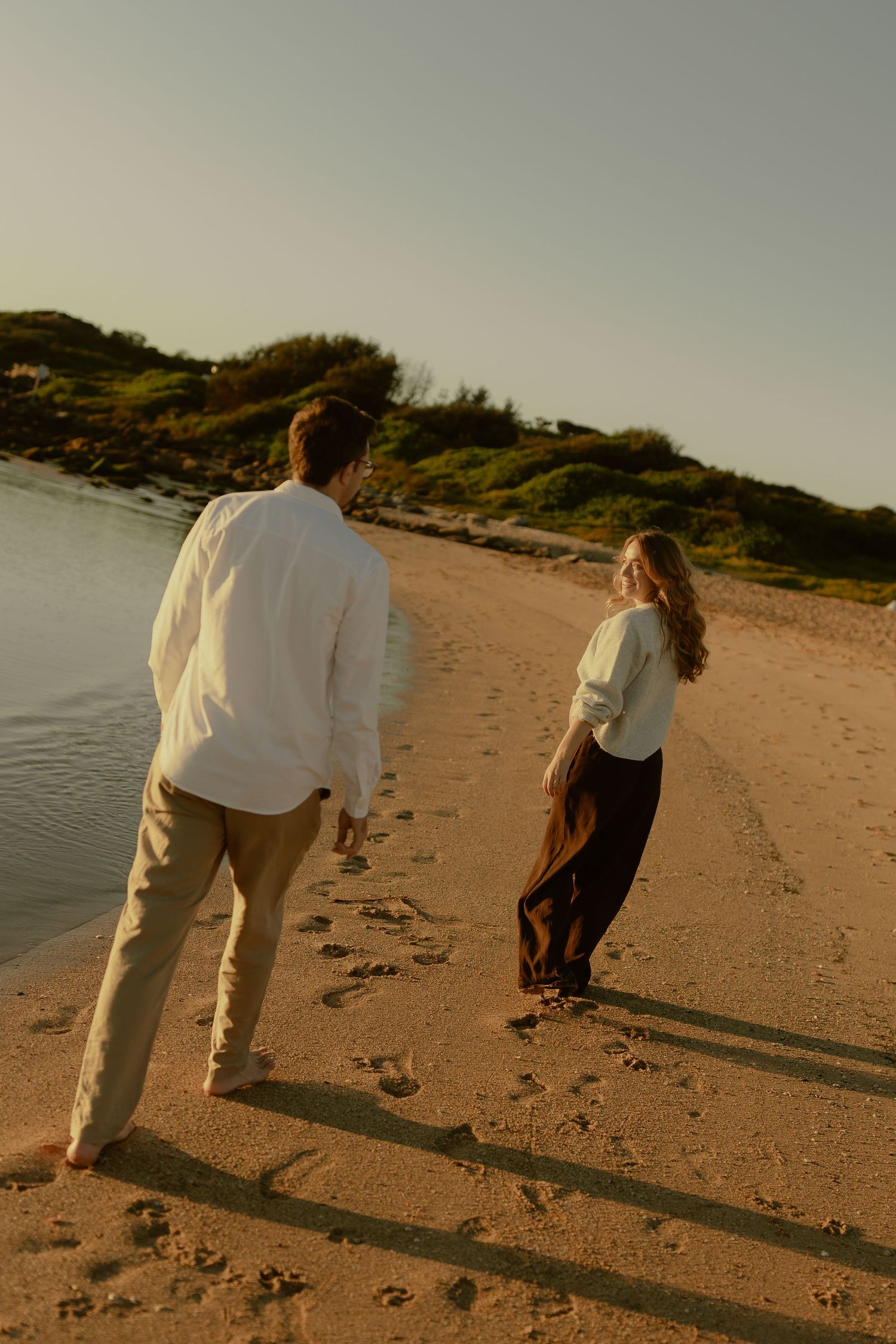 Couple walking barefoot on sandy beach at golden hour, woman in white top and dark skirt looks back smiling.