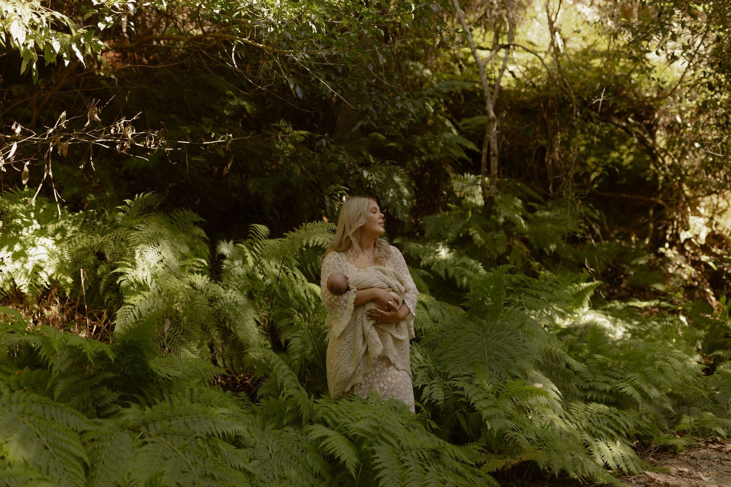 A woman in a flowing dress stands among lush green ferns in a sunlit forest.
