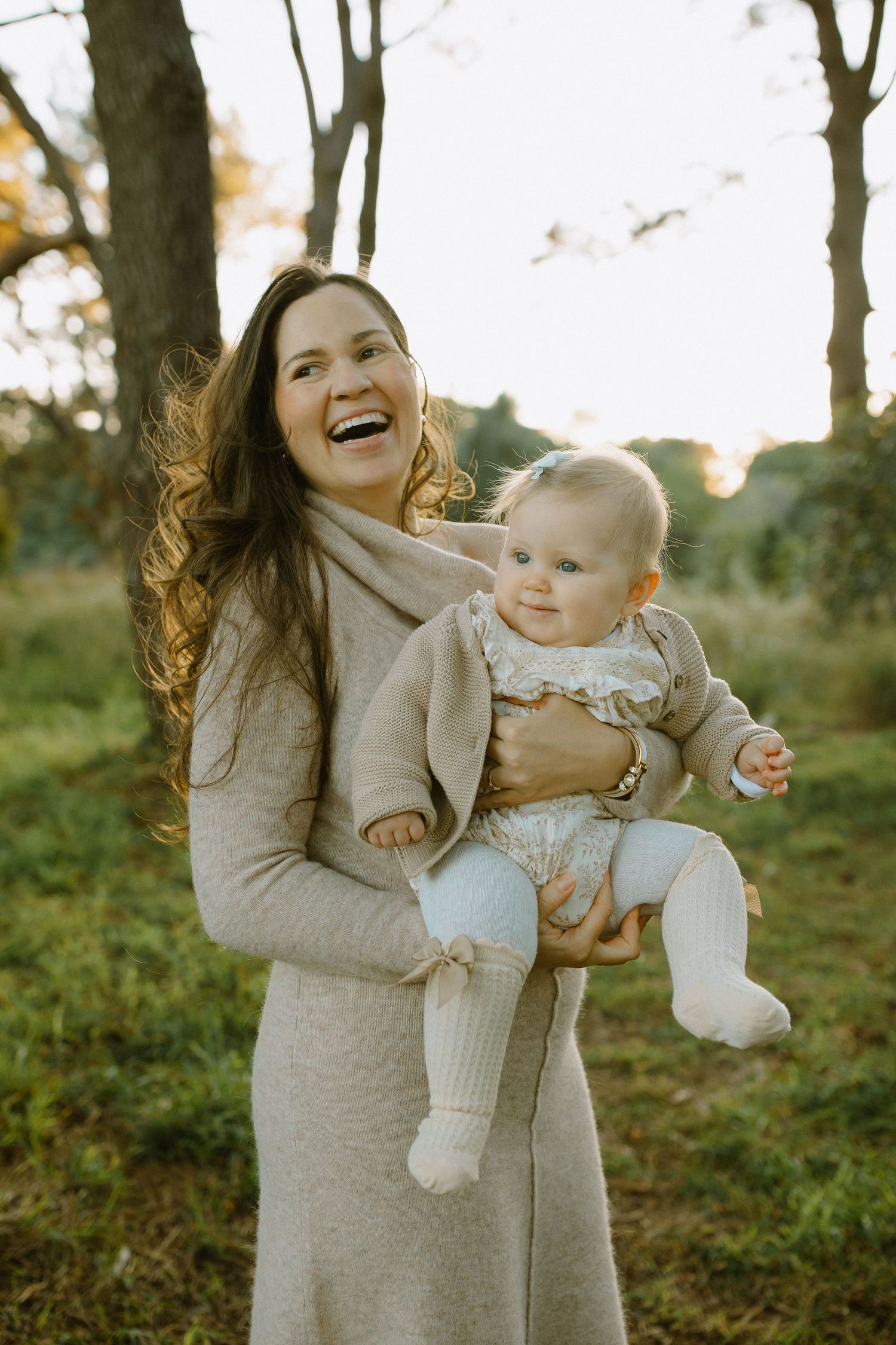 Woman in gray dress laughs joyfully while holding baby in autumn outdoor setting.