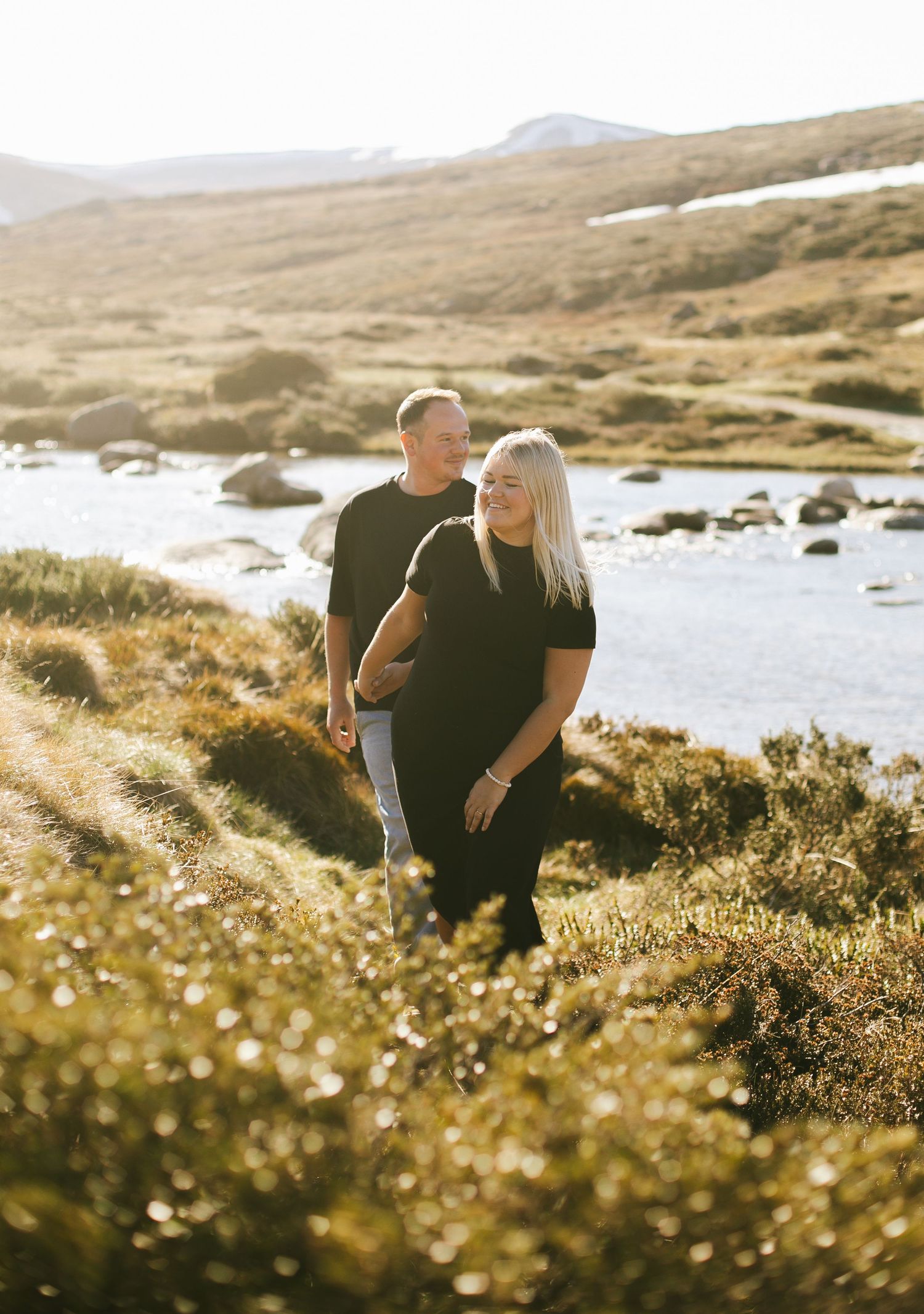 Couple enjoying a peaceful moment together in golden sunlight by a mountain stream with rolling hills in the background.