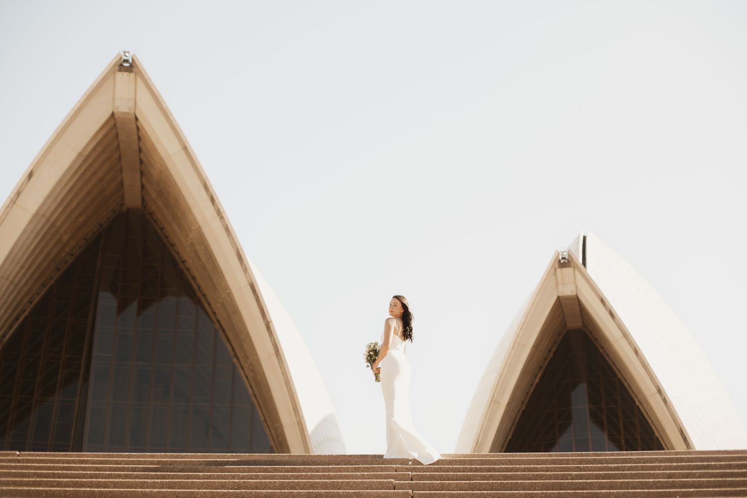 Wide architectural shot of bride standing between Sydney Opera House sails in elegant white dress.