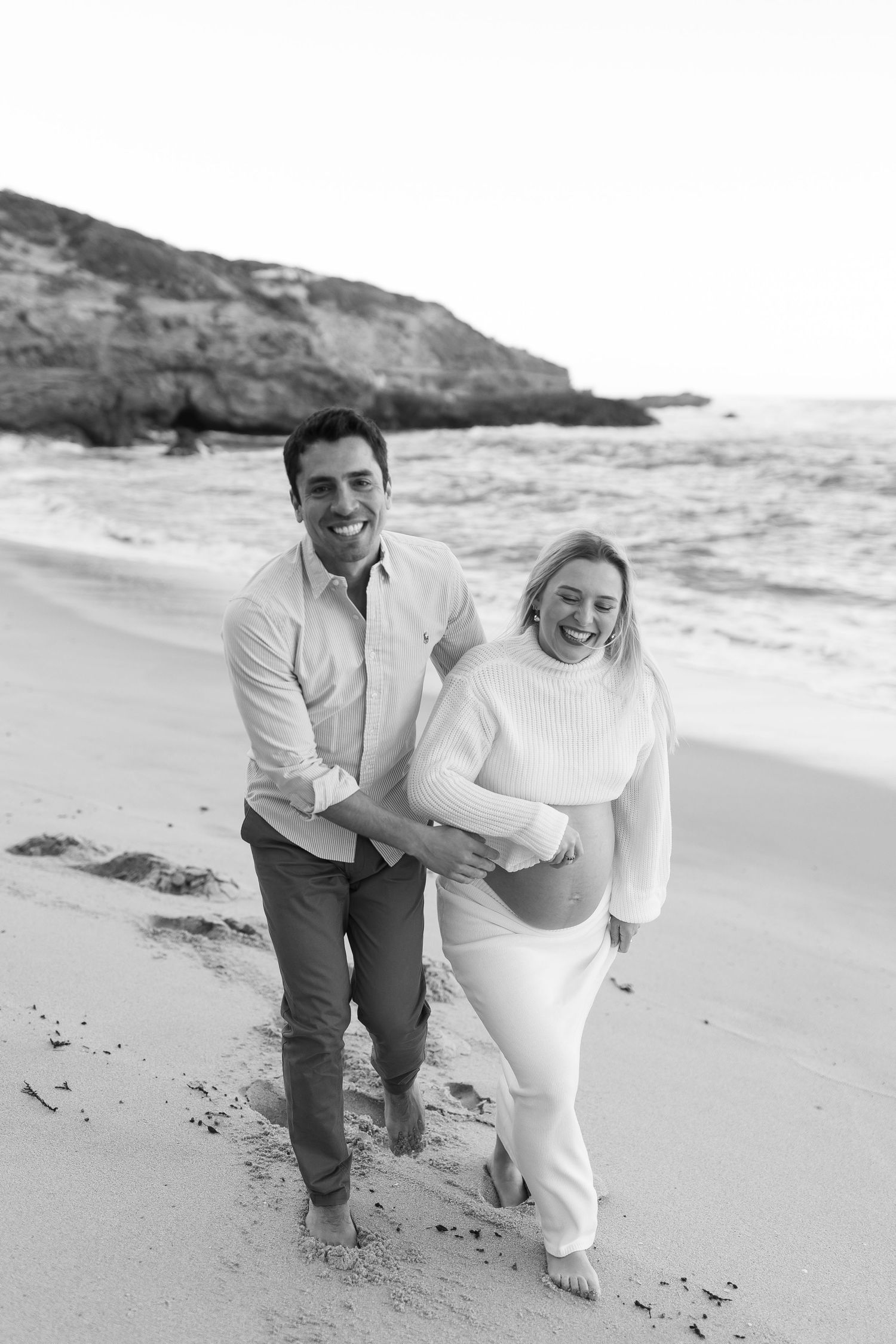 A joyful black and white photo captures an expectant couple laughing while walking along a coastal shoreline.