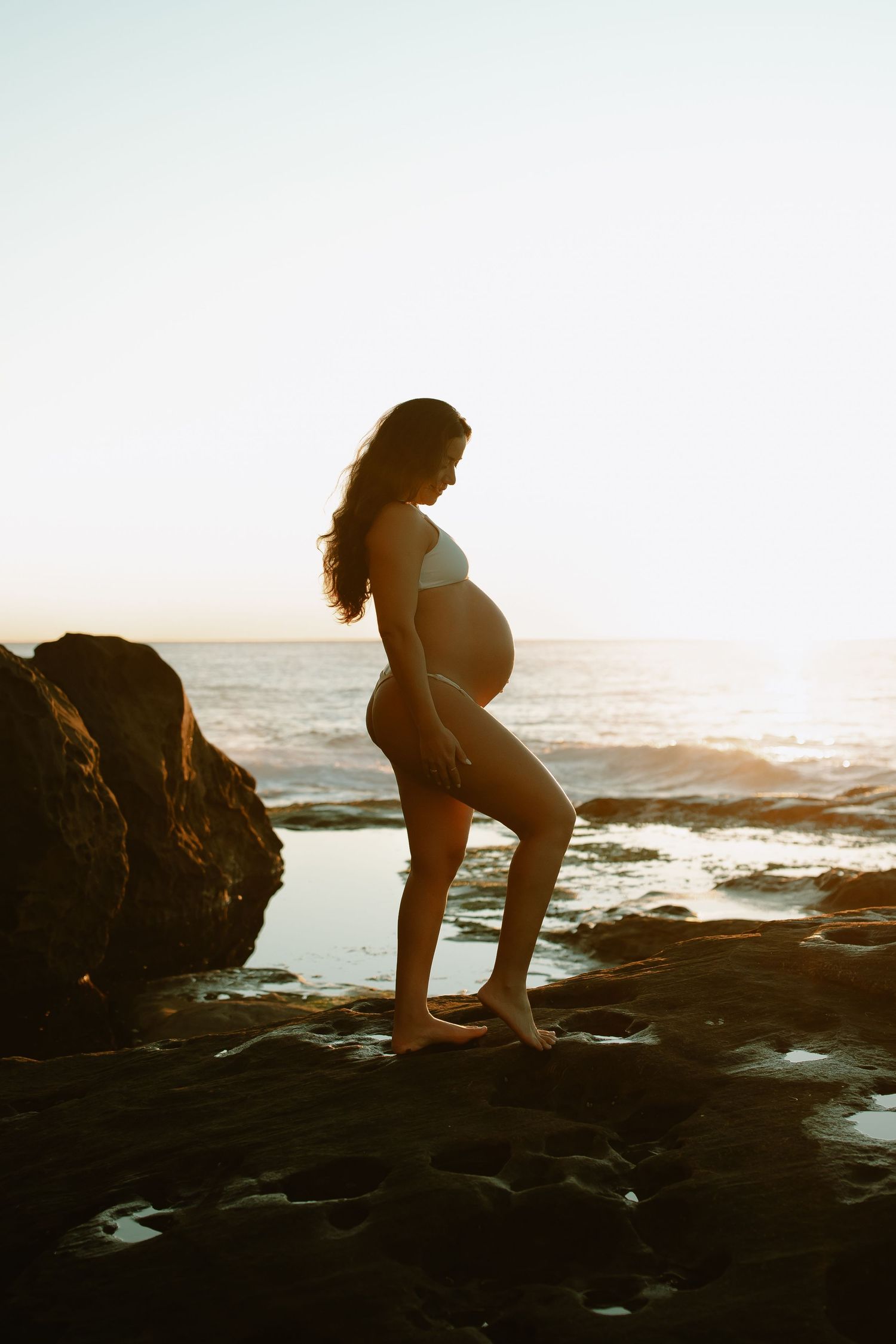 Silhouette of expectant mother standing on rocky beach at sunset with waves and ocean in background.