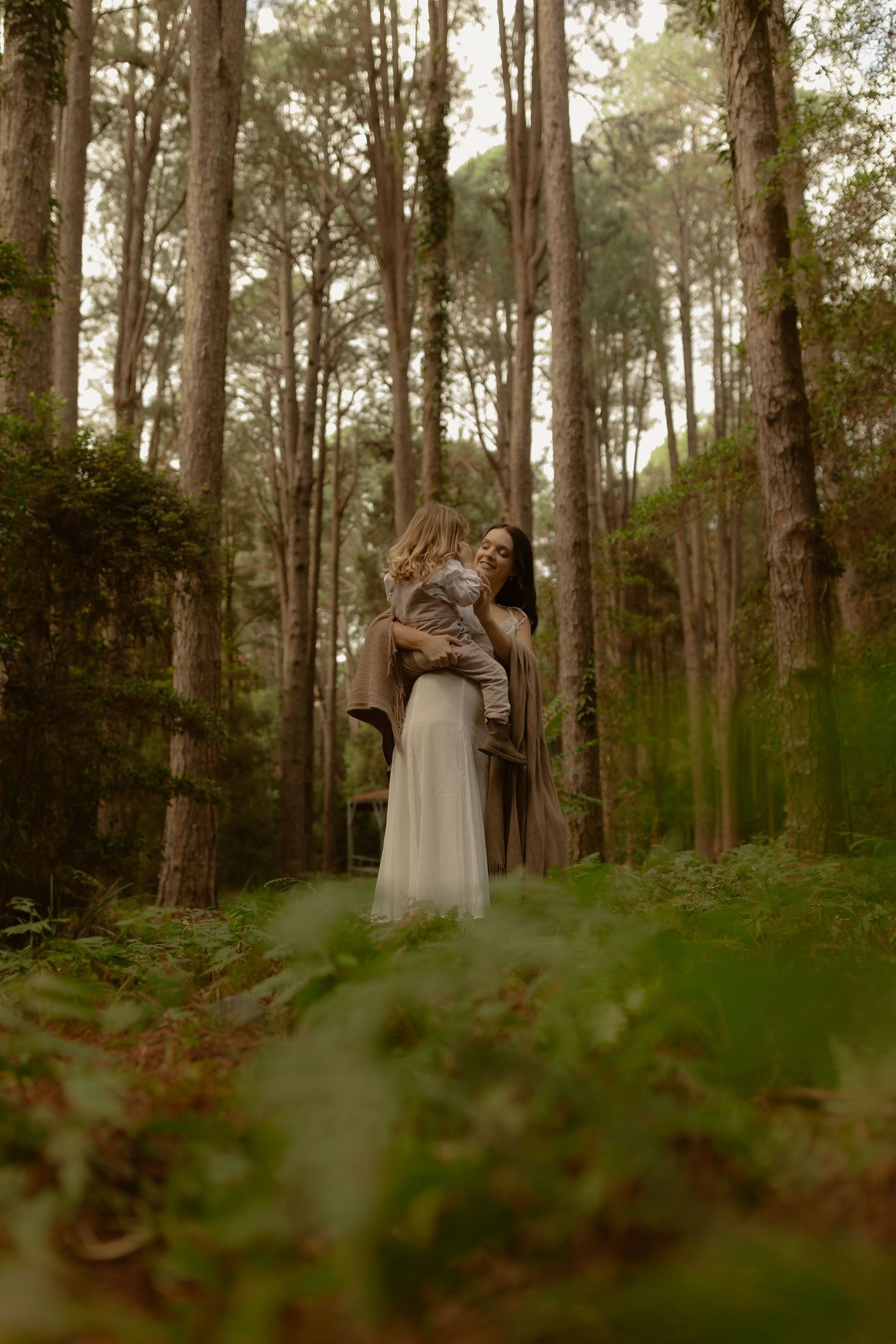 A mother holds her young child in a misty forest, both dressed in earthy tones, surrounded by tall pine trees.
